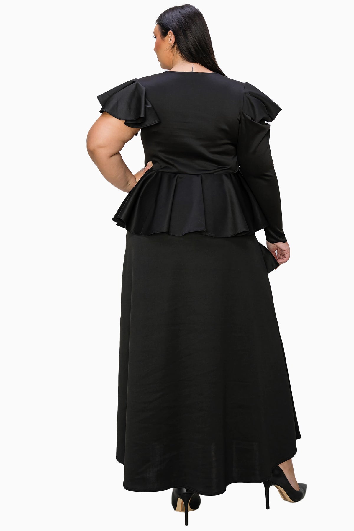 A woman modeling a black two-piece outfit from the back, featuring a long-sleeve peplum top and a matching long skirt, paired with black heels against a plain white background.
