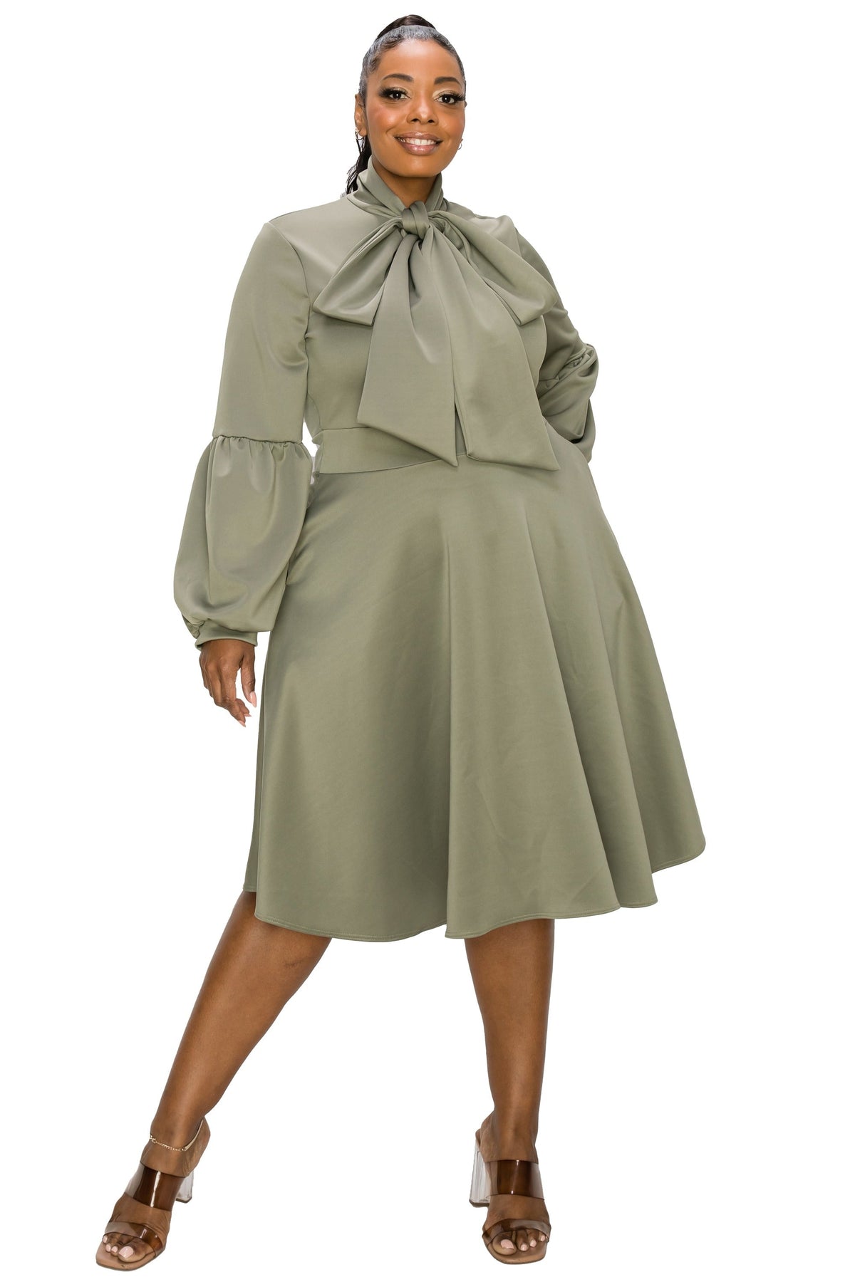 A woman modeling a sage green midi dress with a high neck, large bow detail, and voluminous long sleeves, paired with brown heeled sandals against a plain white background.