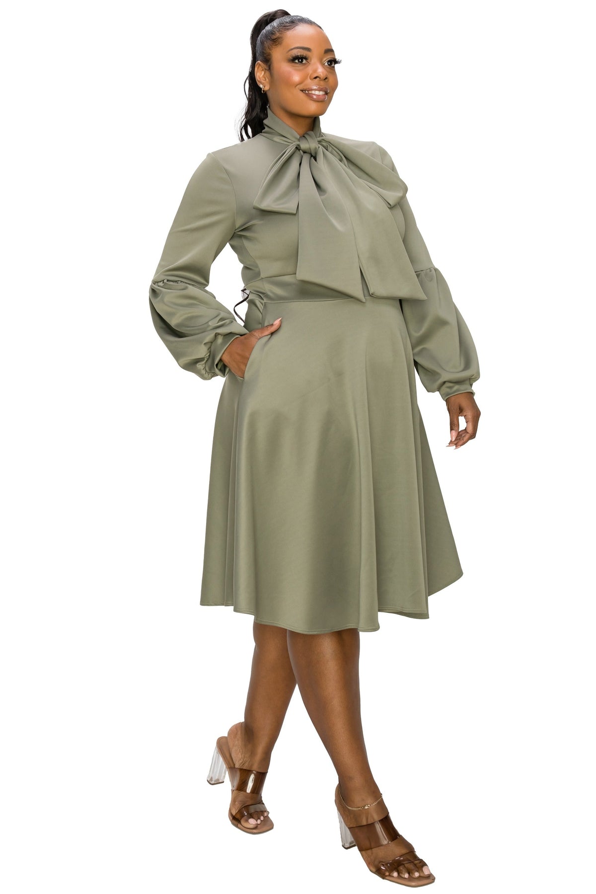 A woman modeling a sage green midi dress with a large ruffled bow tie neckline, long sleeves, and a flared skirt, paired with metallic brown high-heeled sandals against a plain white background.