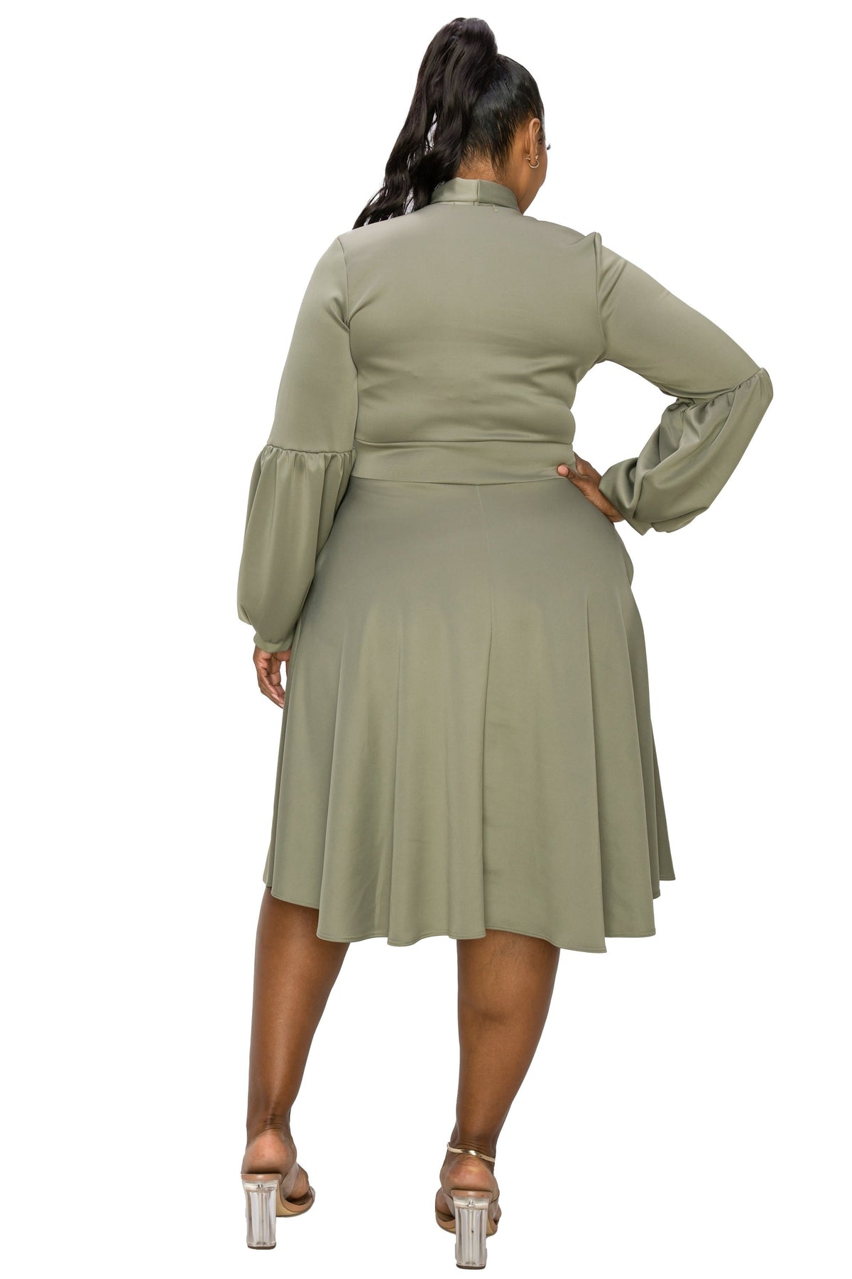 A woman shown from the back wearing a sage green long-sleeve midi dress with a high collar and pleated skirt, paired with clear high-heeled sandals, against a plain white background.
