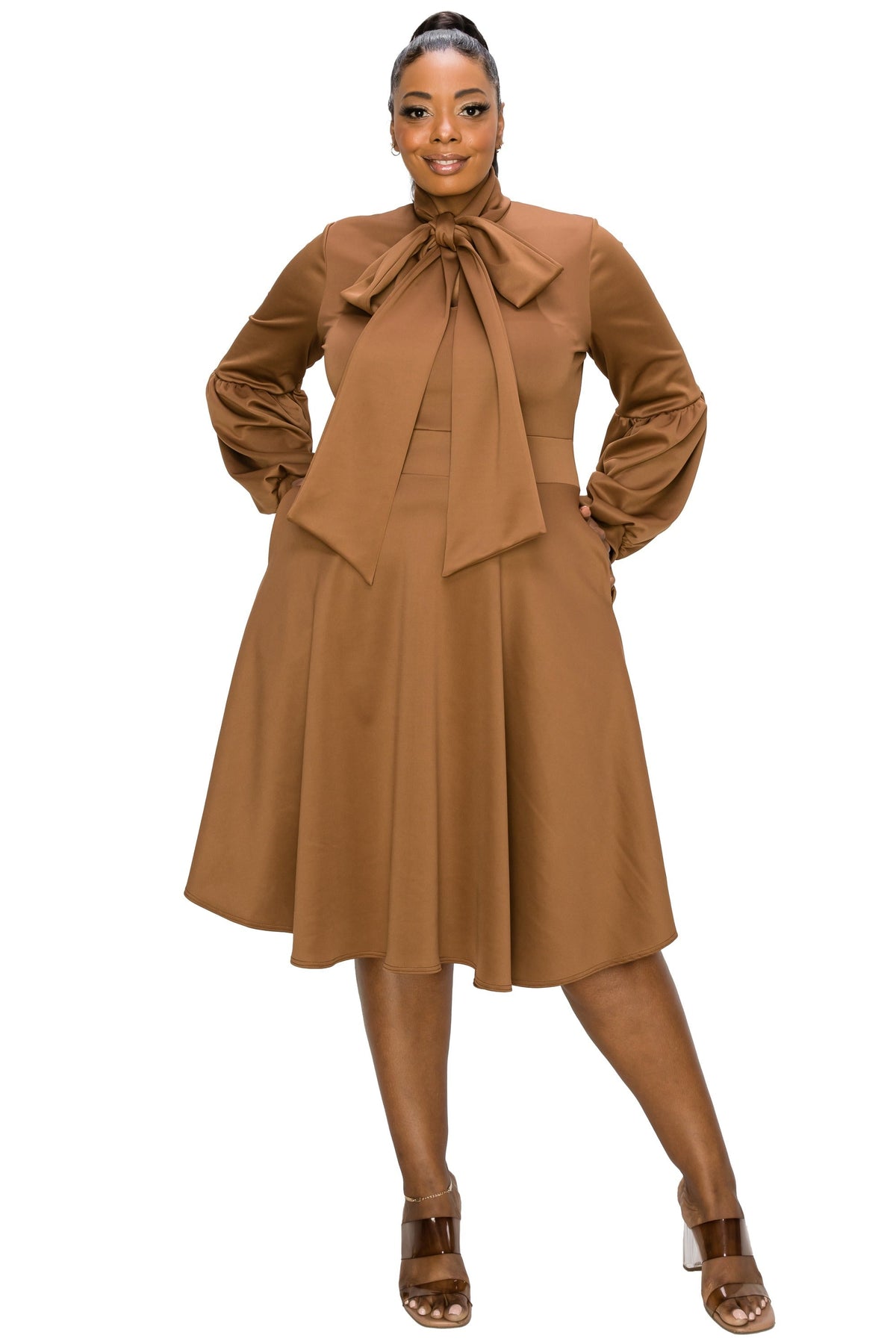 A woman modeling a brown long-sleeve midi dress with a large bow tie at the collar, side pockets, and a flared skirt, paired with brown heeled sandals against a plain white background.