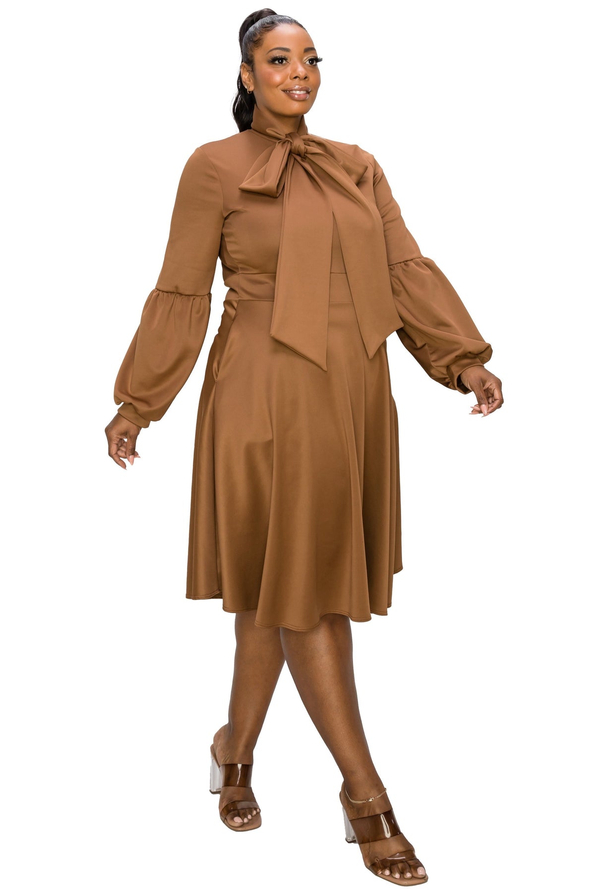 A woman modeling a brown midi dress with a high neck, large bow detail, and voluminous long sleeves, paired with brown heeled sandals against a plain white background.