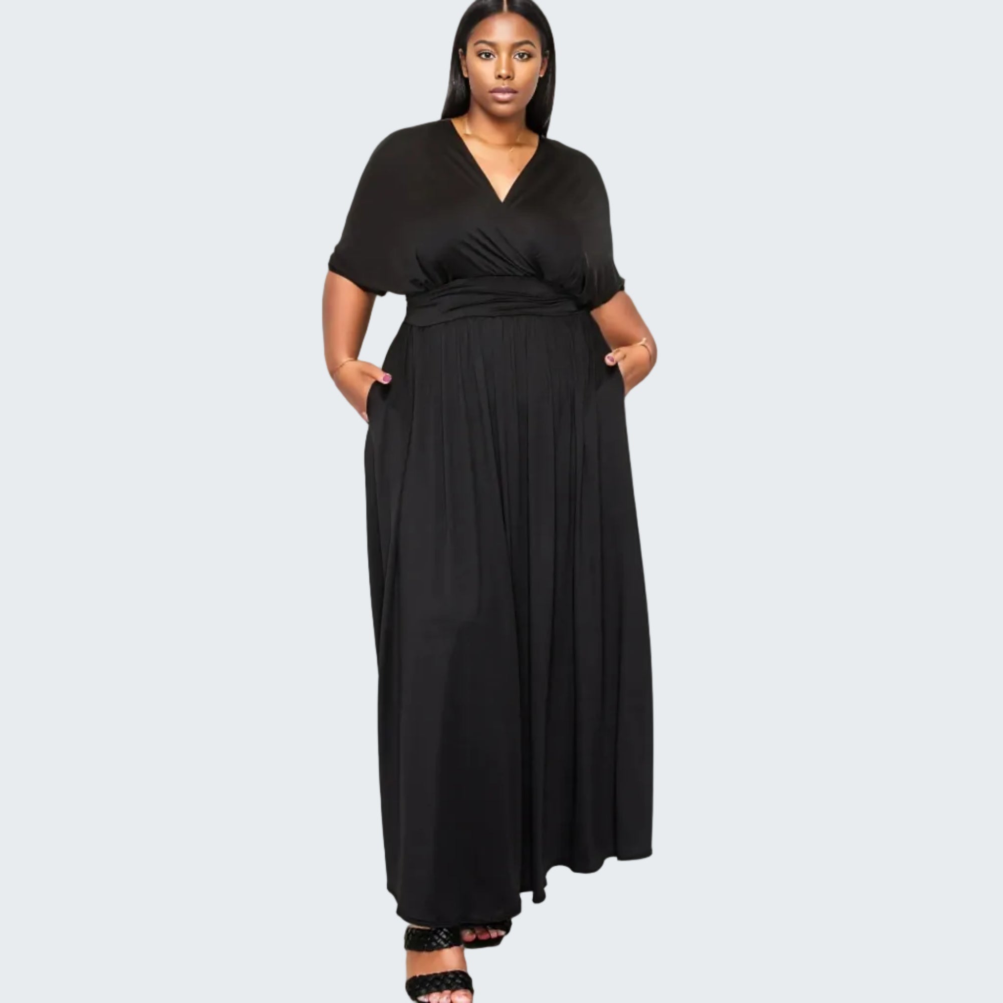 A woman modeling a black maxi dress with short sleeves, a V-neckline, and side pockets, standing against a plain white background.