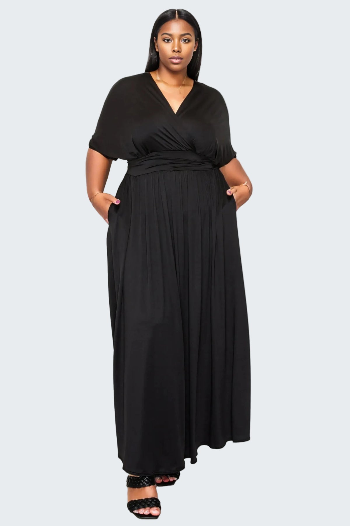 A woman modeling a black maxi dress with short sleeves, a V-neckline, and side pockets, standing against a plain white background.