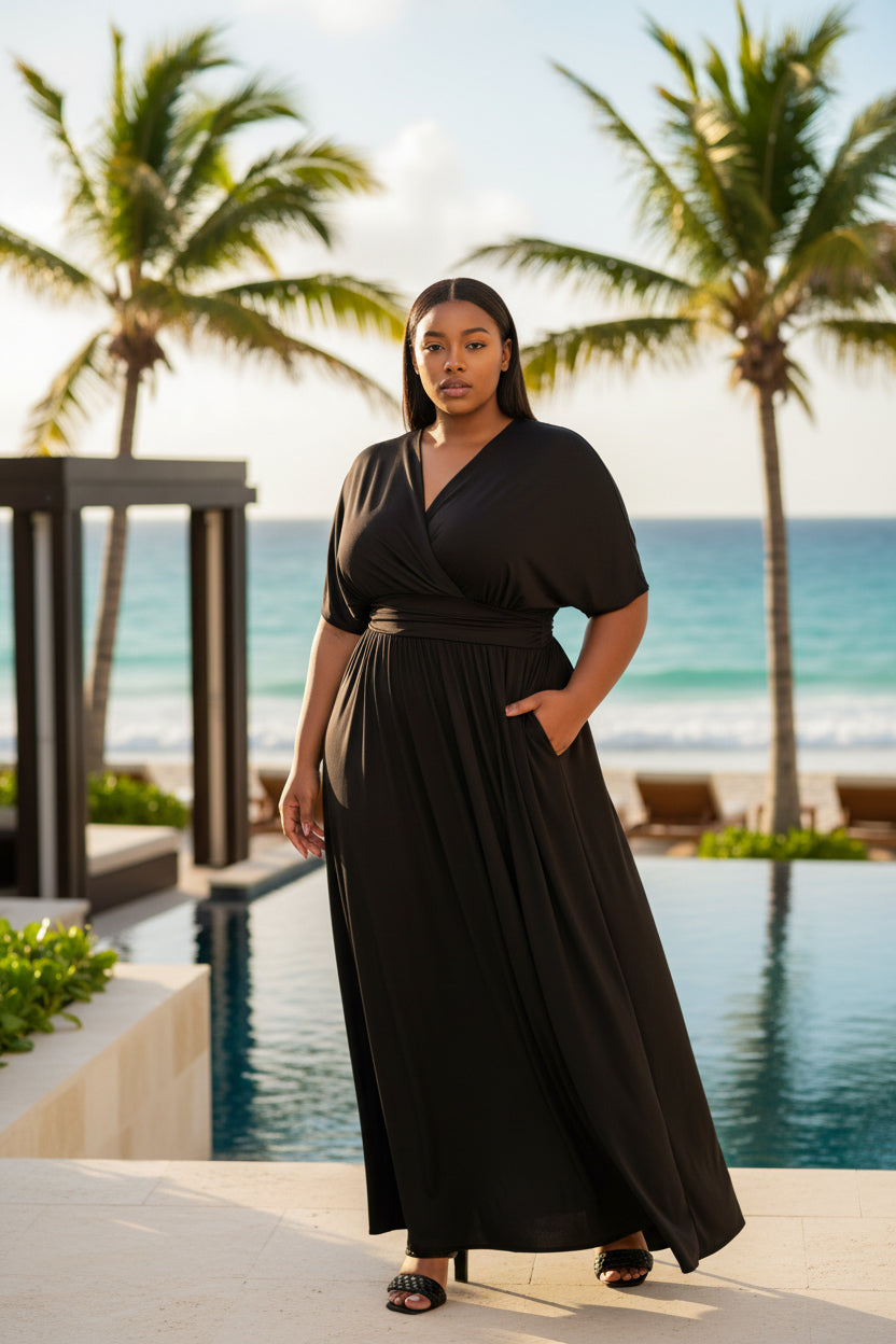 A woman modeling a black maxi wrap dress with short sleeves, a V-neckline, and side pockets, paired with black heeled sandals against a plain white background.