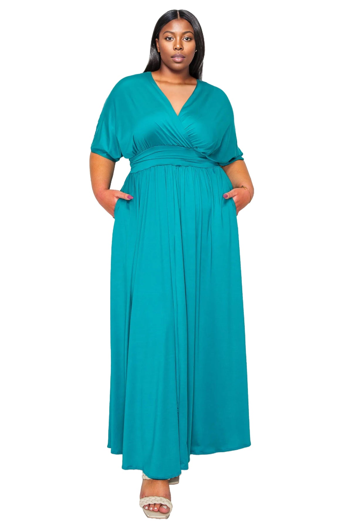 A woman modeling a teal green maxi dress with short sleeves, a V-neckline, and side pockets, standing against a plain white background.