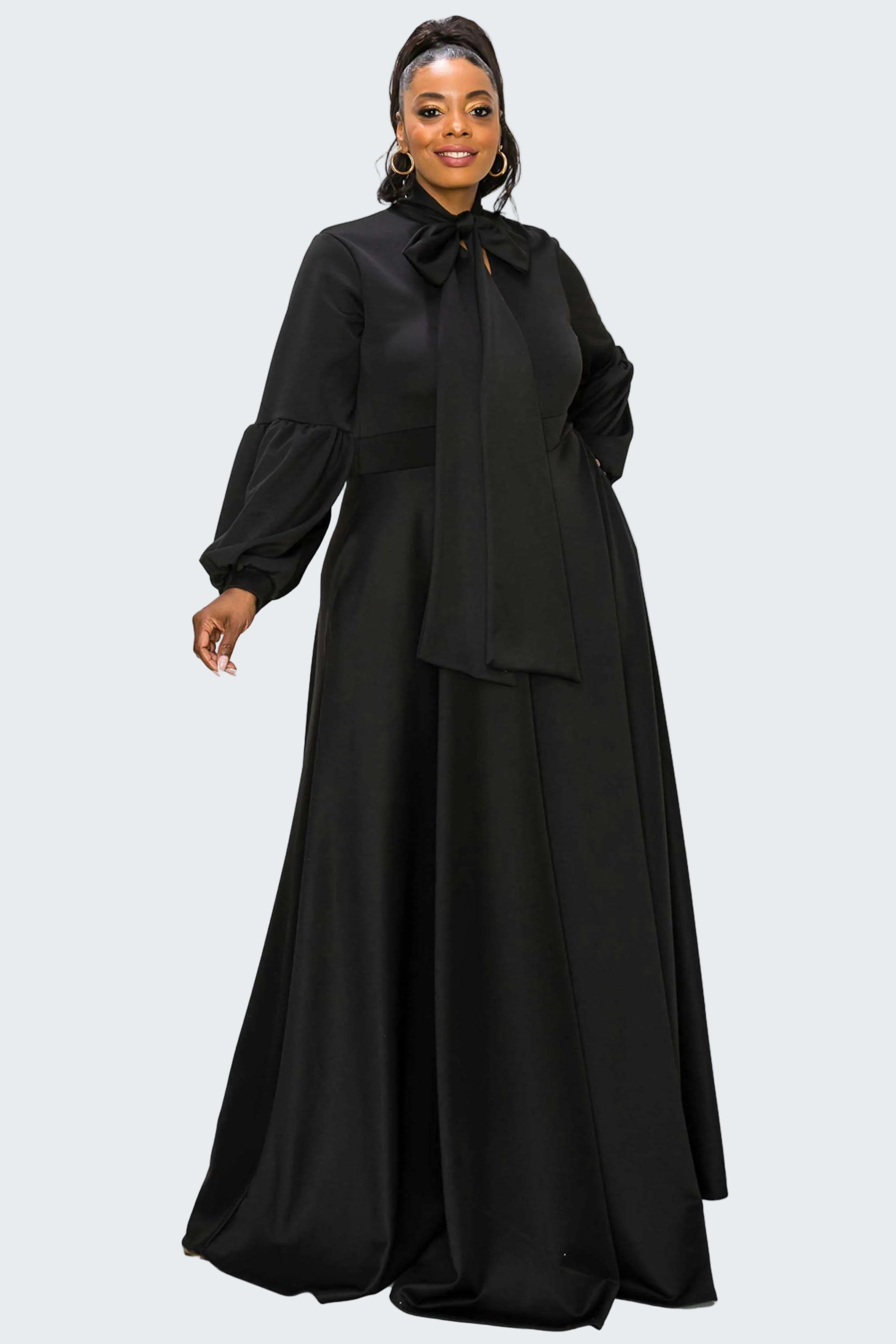 A woman modeling a long black dress with a high neck, large bow at the collar, long balloon sleeves, and a flowing skirt, photographed against a plain white background.