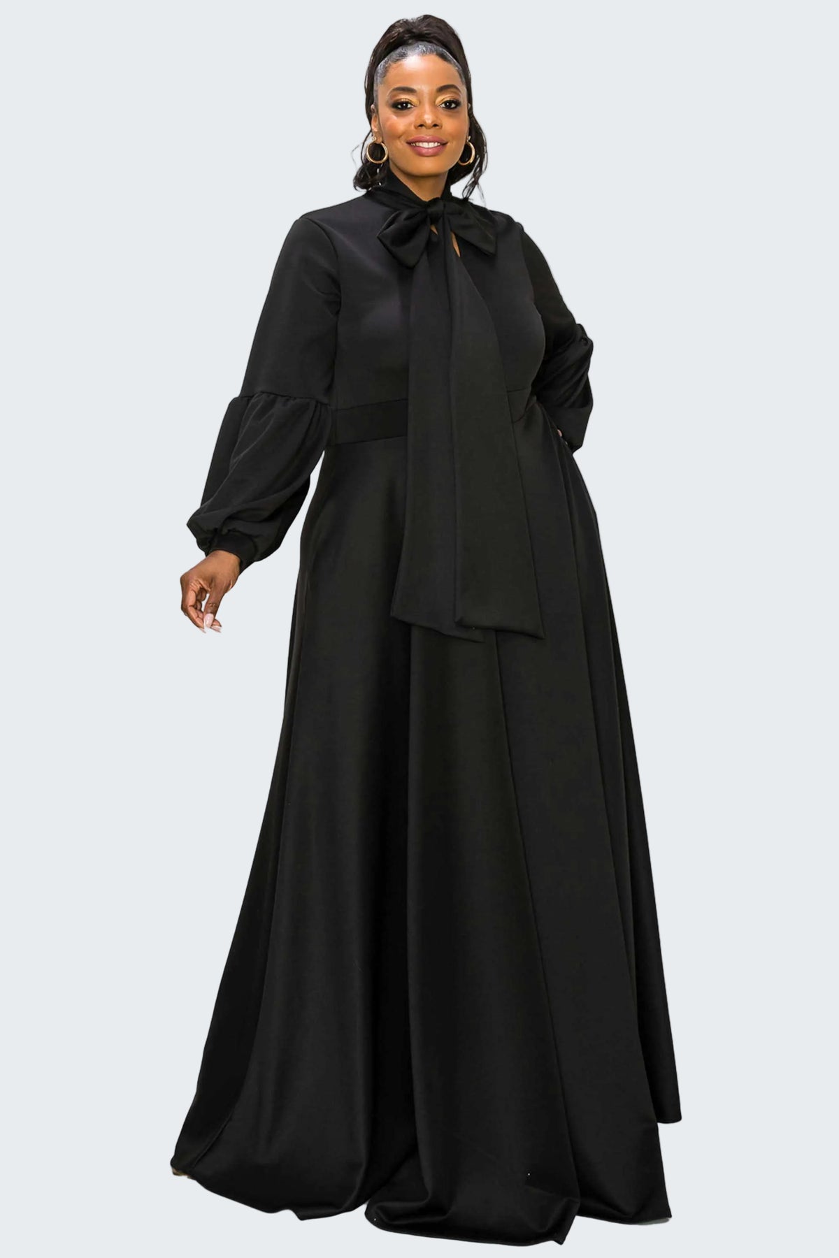 A woman modeling a long black dress with a high neck, large bow at the collar, long balloon sleeves, and a flowing skirt, photographed against a plain white background.