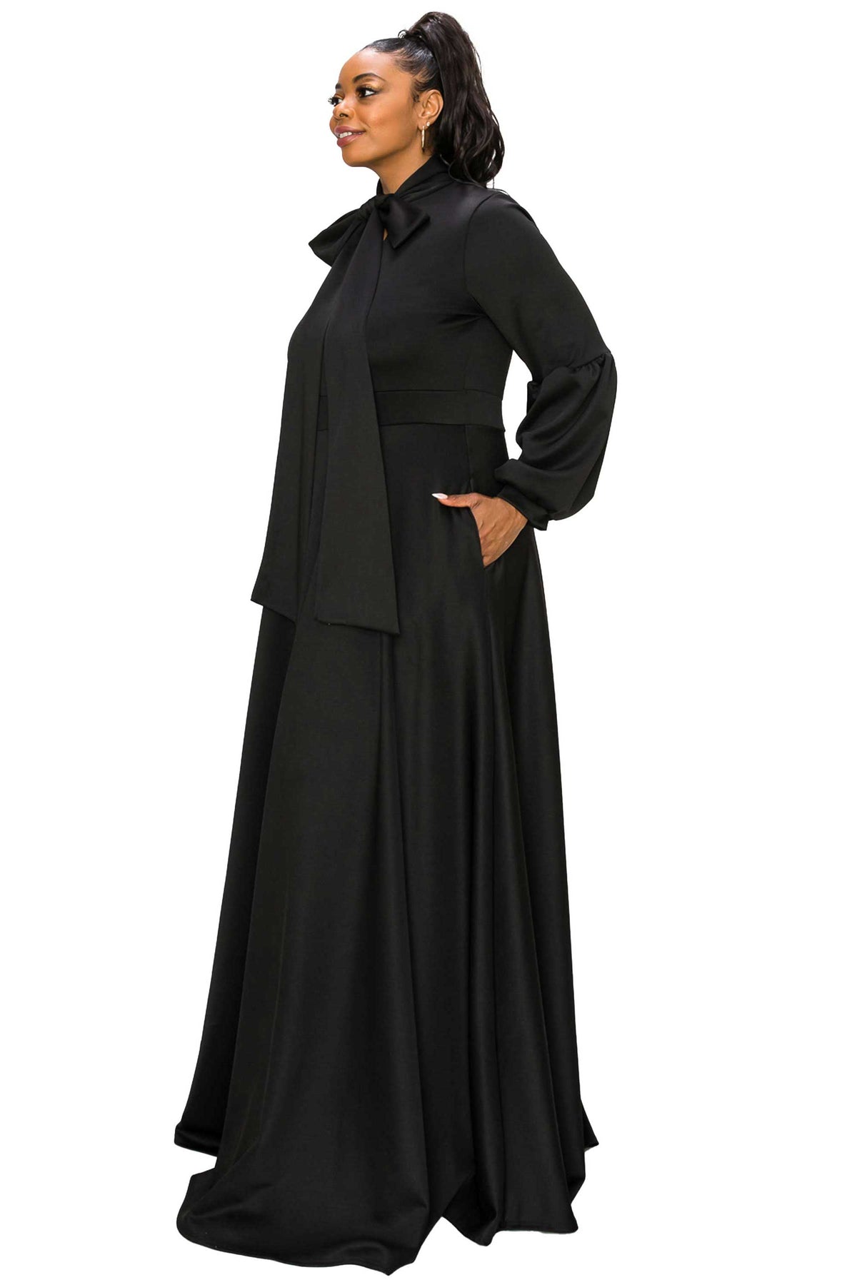 A woman modeling a long black dress with a high collar, long sleeves, and a draped overlay, shown against a plain white background.