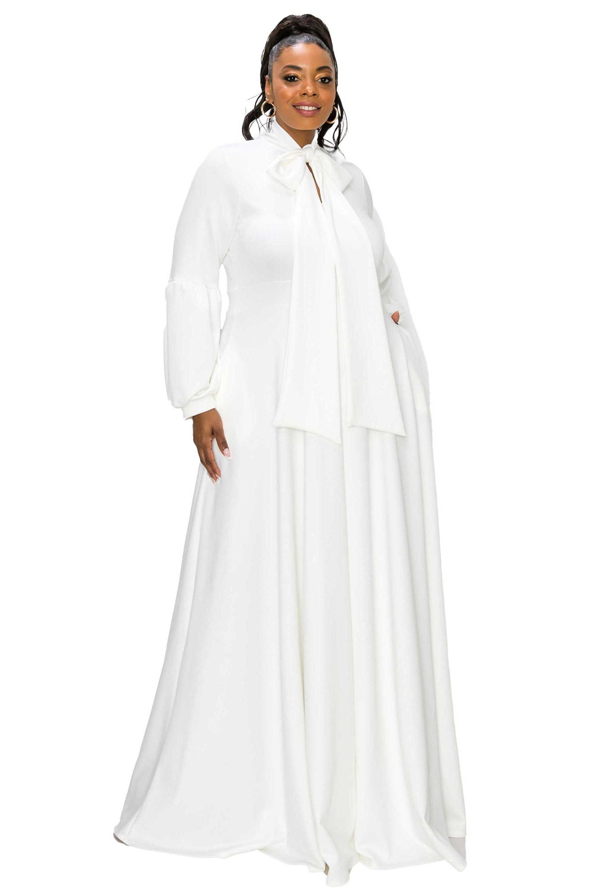 A woman modeling a long white flowing dress with a high collar, bow at the neckline, and voluminous long sleeves against a plain white background.