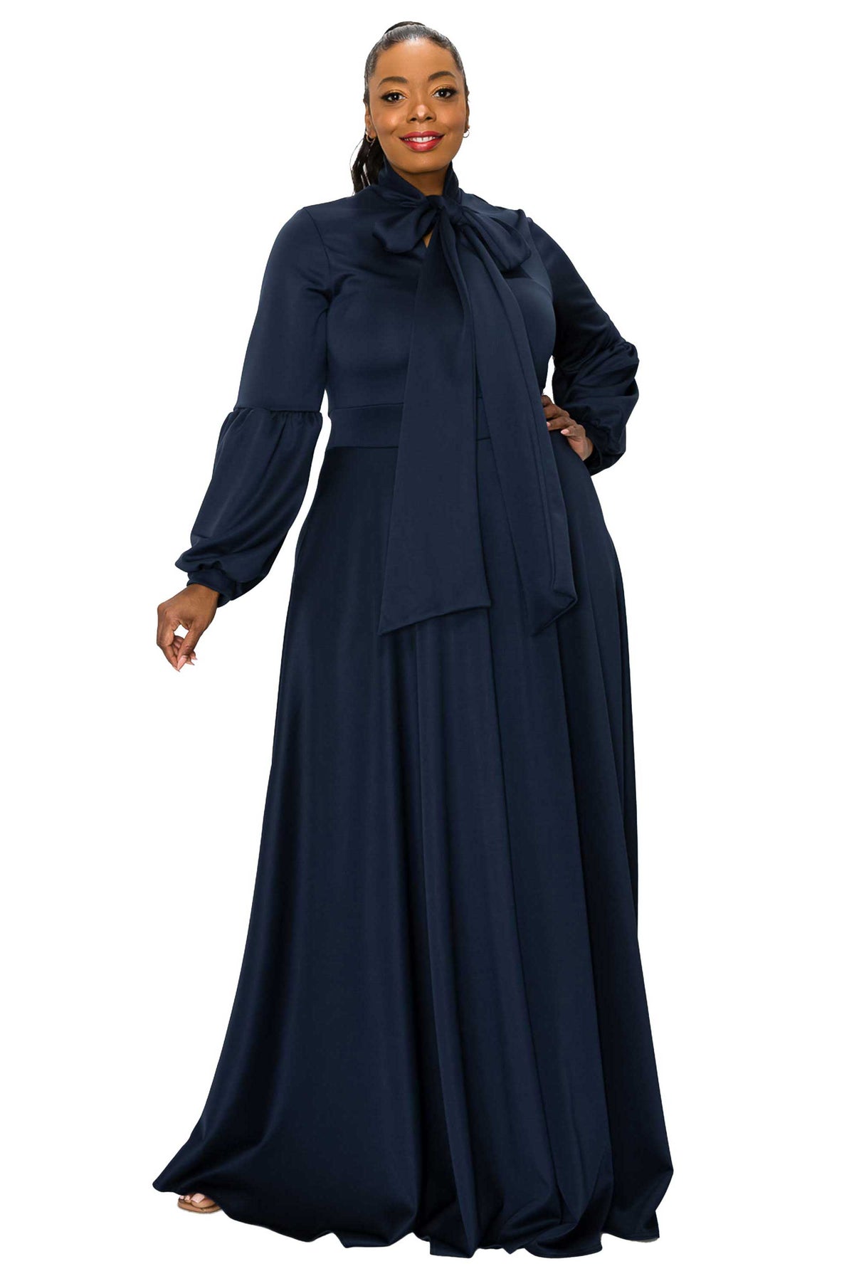 A woman modeling a long navy blue maxi dress with a high neck, large bow detail at the collar, long sleeves, and a flowing skirt, set against a plain white background.