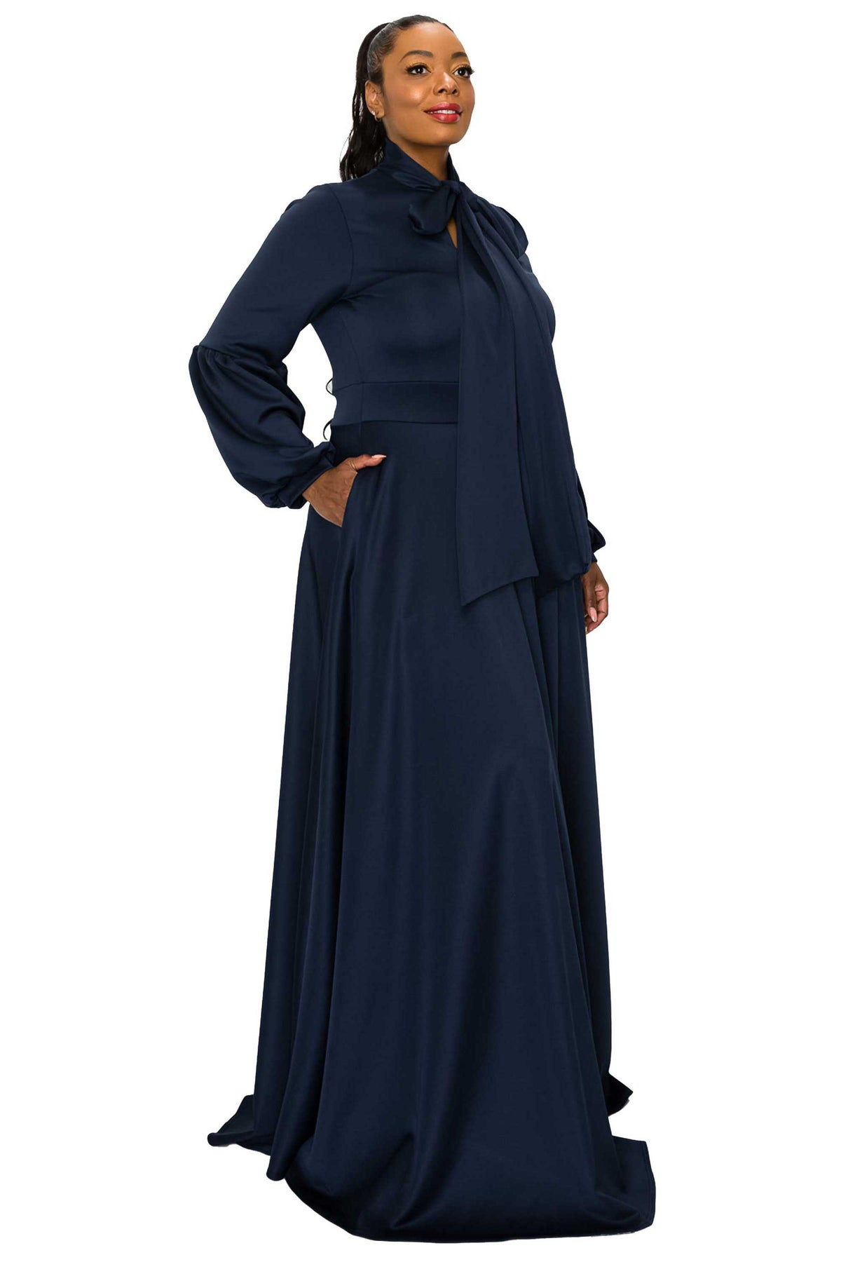 A woman modeling a long navy blue maxi dress with a high neck, bow detail at the collar, long sleeves, and side pockets, set against a plain white background.
