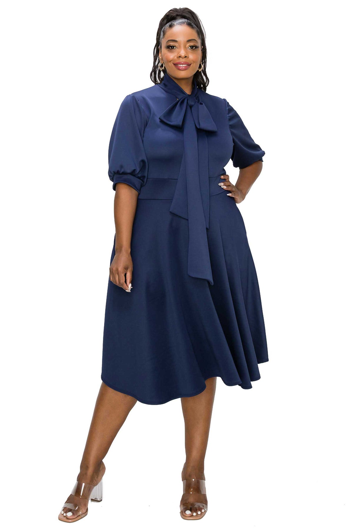 A woman modeling a navy blue midi dress with a high neck, large bow detail at the collar, puff sleeves, and a flared skirt, paired with clear high-heeled sandals against a plain white background.