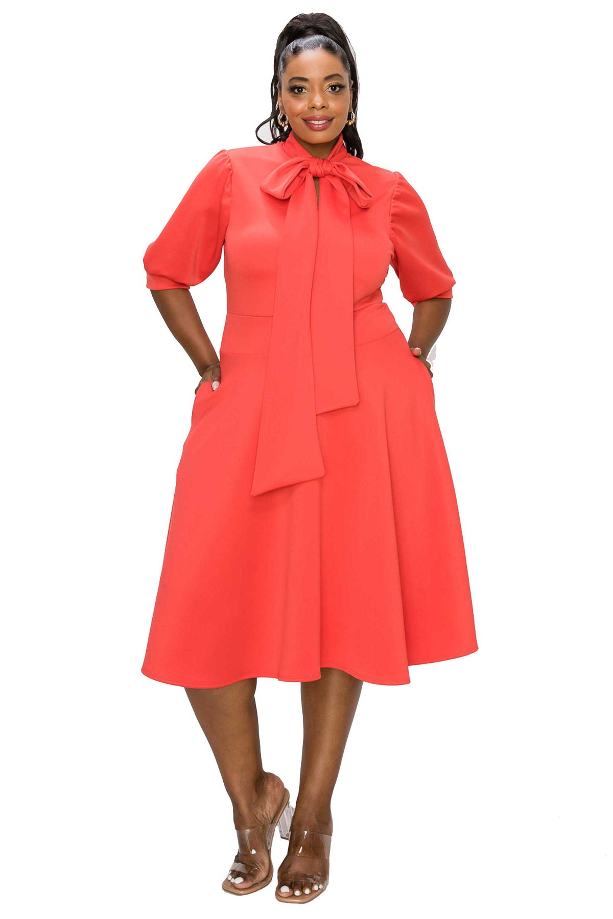 A woman modeling a bright coral red midi dress with puff sleeves, a large bow at the collar, and side pockets, paired with clear high-heeled sandals against a plain white background.