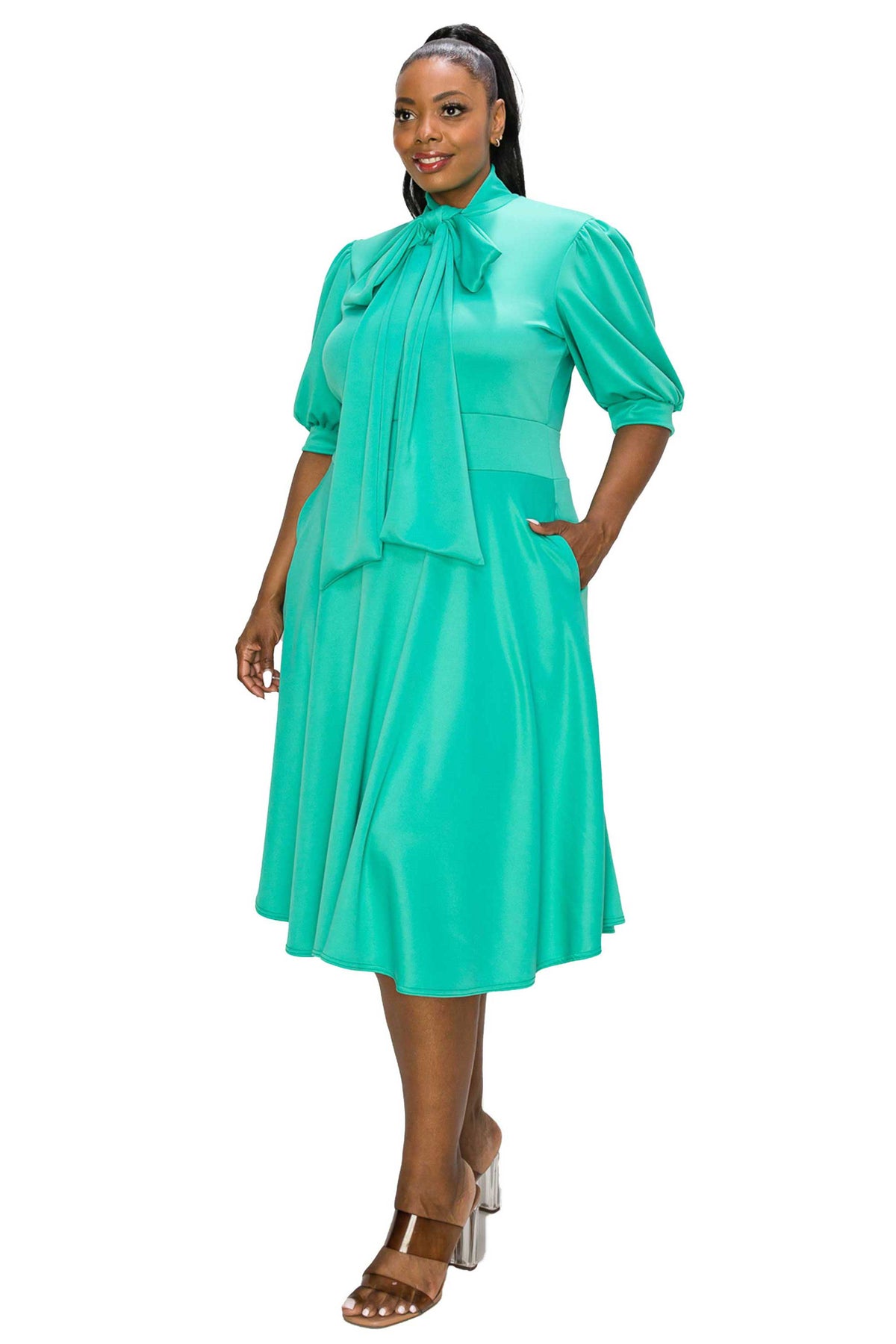 A woman modeling a bright turquoise green midi dress with puff sleeves, a high neck, and a large bow detail at the collar, paired with metallic brown high-heeled sandals against a plain white background.