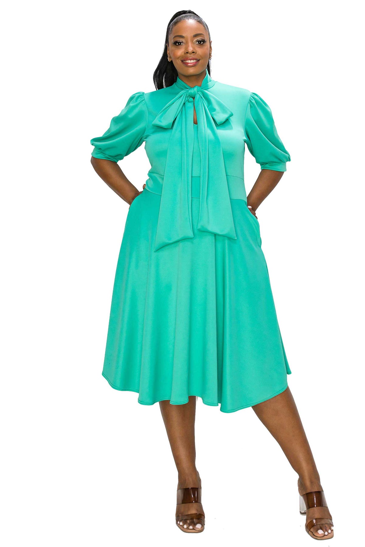 A woman modeling a bright turquoise green midi dress with puff sleeves, a high neck, and a large bow detail at the collar, paired with brown heeled sandals against a plain white background.