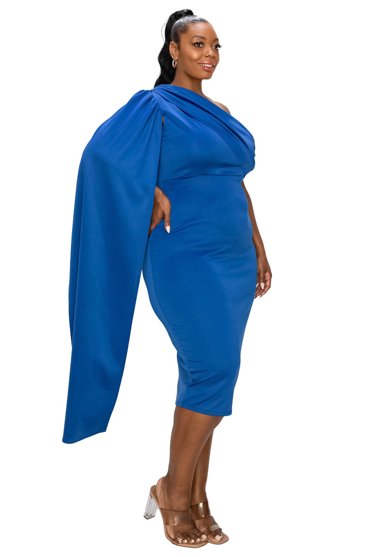 A woman modeling a bright royal blue one-shoulder midi dress with a dramatic draped capelet detail, paired with brown heeled sandals, against a plain white background.