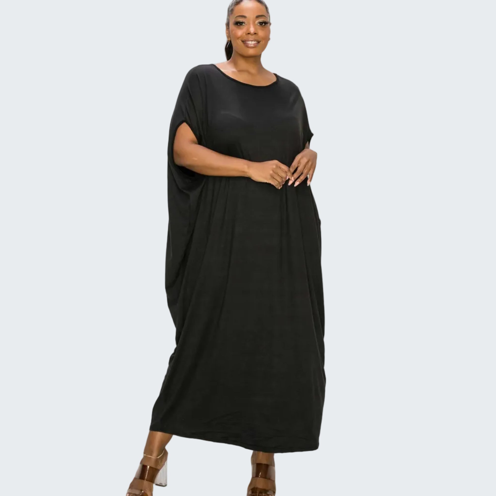 A woman modeling a black, loose-fitting maxi dress with short draped sleeves and side pockets, paired with brown block-heel sandals, against a plain white background.