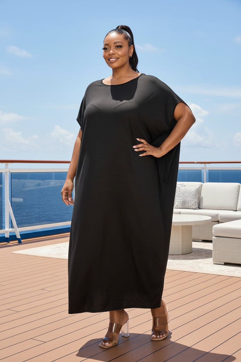 A woman modeling a loose, black, midi-length shift dress with short sleeves and a boat neckline, paired with brown high-heeled sandals, against a plain white background.