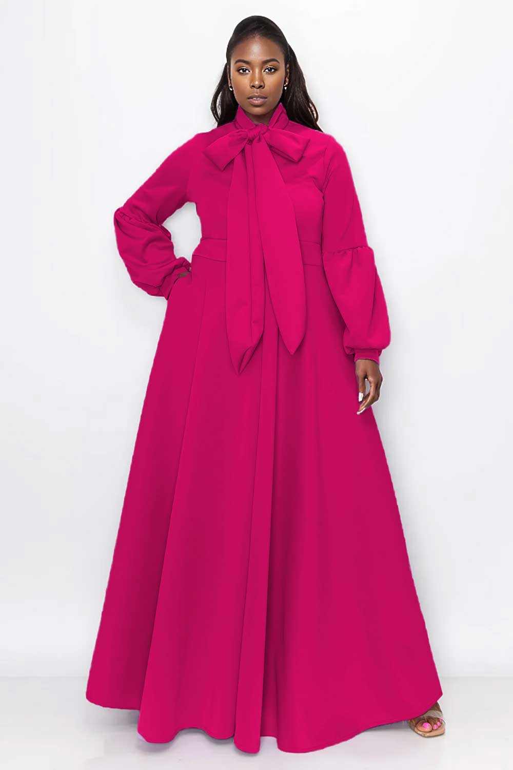 A woman modeling a bright magenta maxi dress with long balloon sleeves, a high mock neck, and a large bow at the collar, set against a plain white studio background.