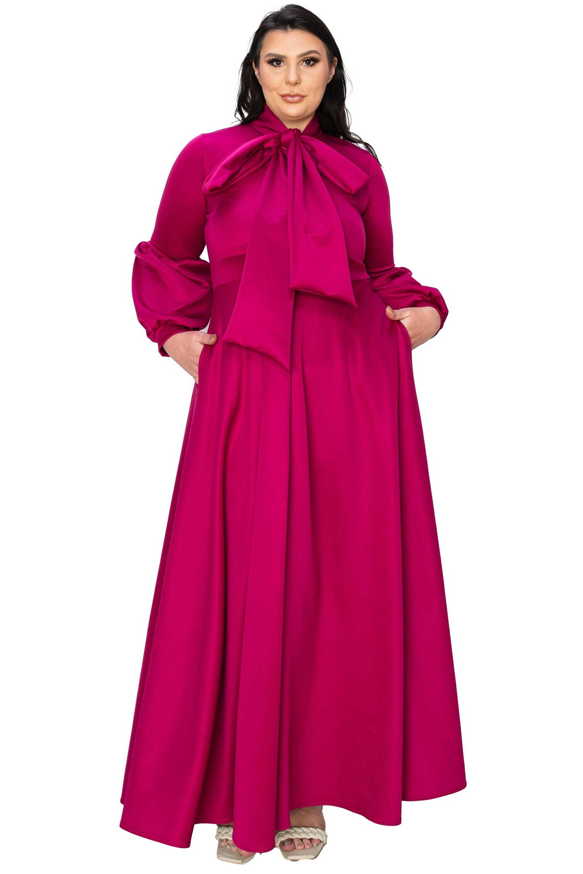 A woman modeling a long magenta maxi dress with a large bow at the neckline, long balloon sleeves, and a flowing skirt, shown against a plain white background.