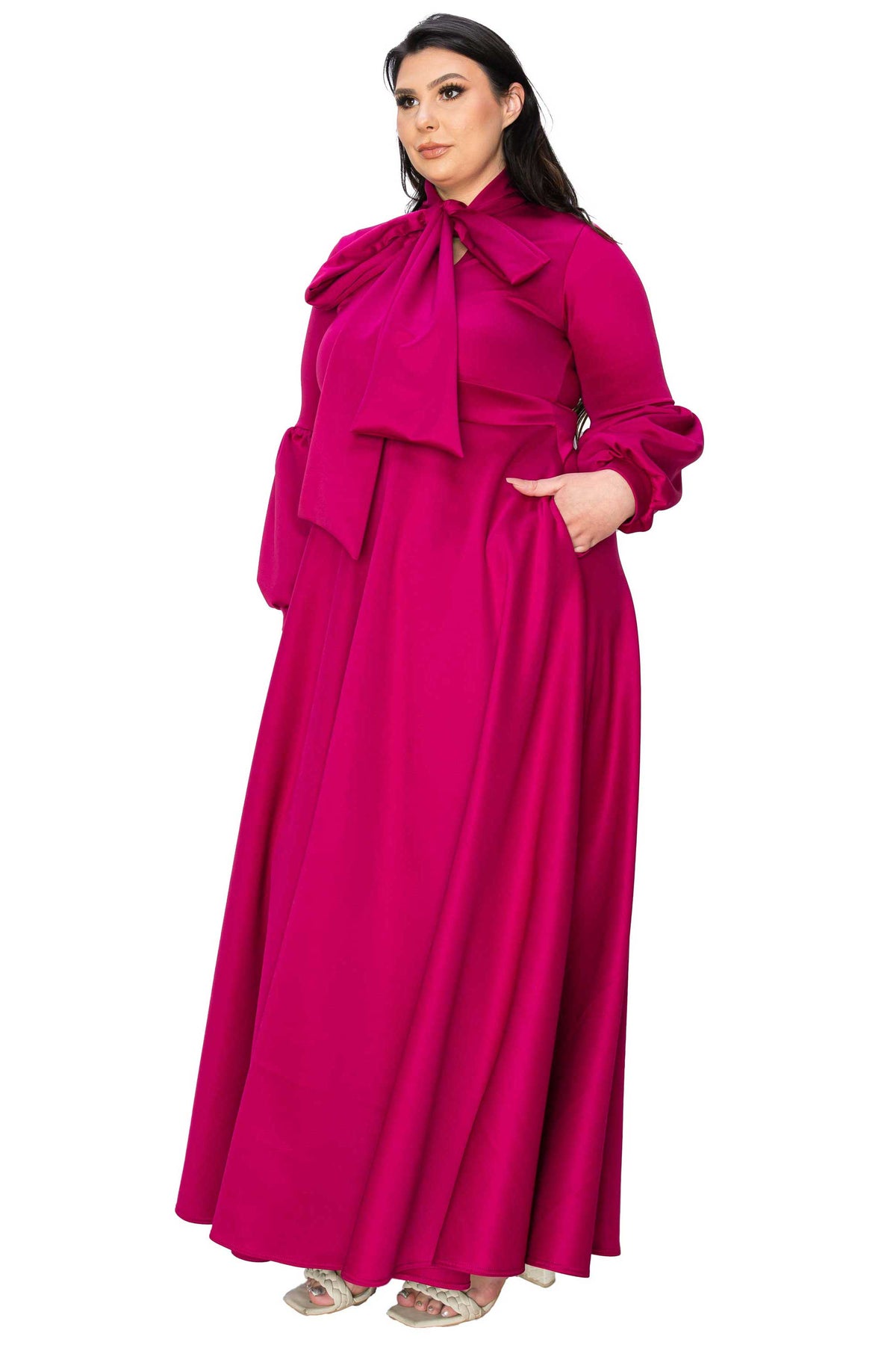 A woman modeling a long magenta maxi dress with a large bow at the collar, long sleeves, and side pockets, shown against a plain white background.