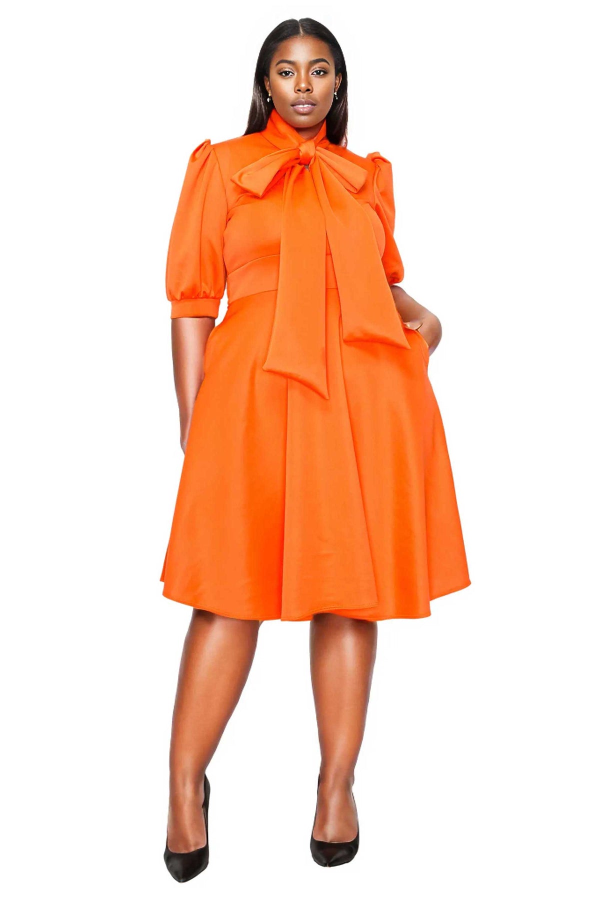 A woman modeling a bright orange midi dress with puff sleeves, a large bow at the high neckline, and a flared skirt, paired with black pointed-toe heels against a plain white background.