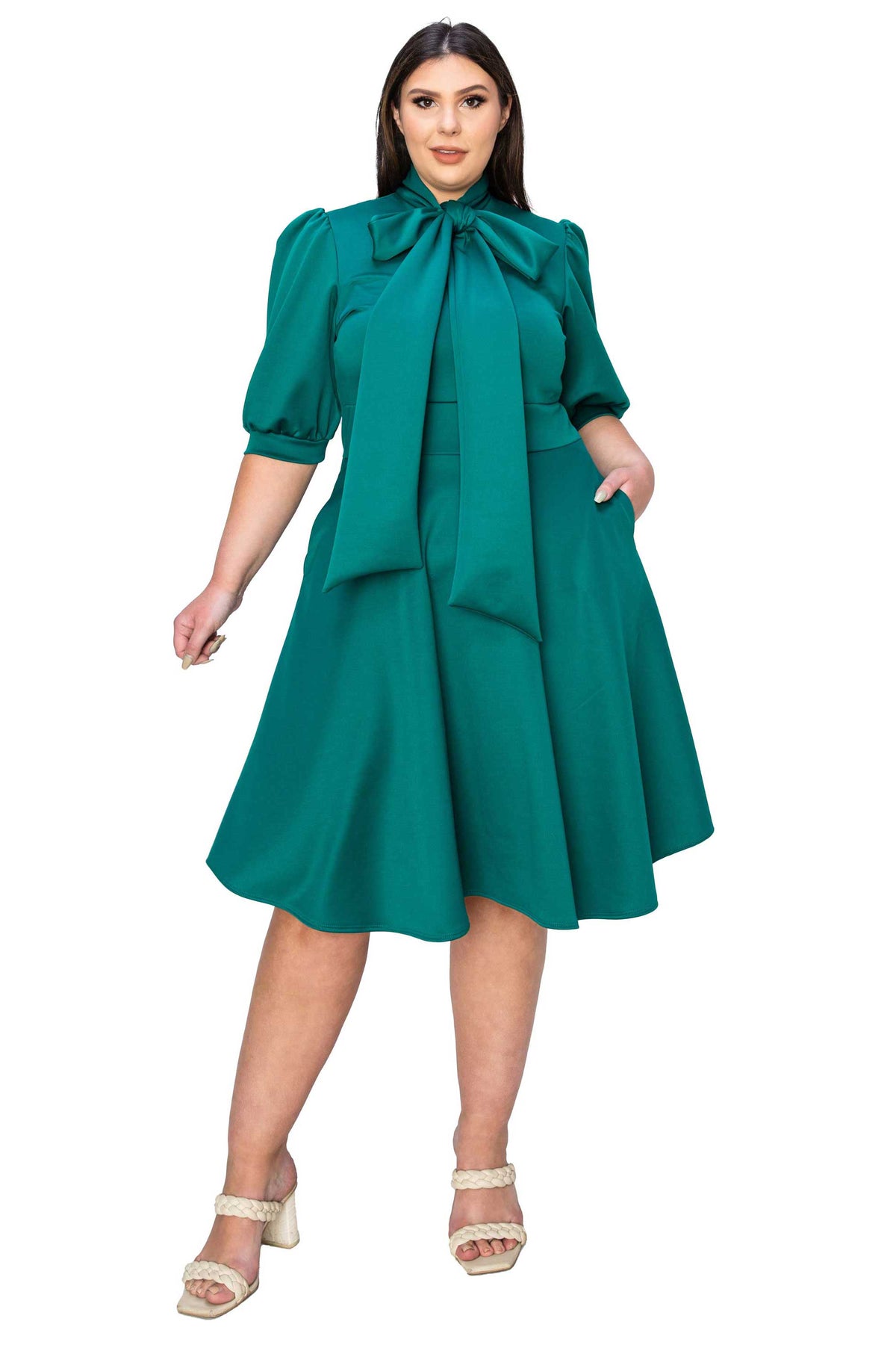 A woman modeling a teal green midi dress with puff sleeves, a large bow at the collar, and a flared skirt, paired with beige strappy high-heeled sandals against a plain white background.