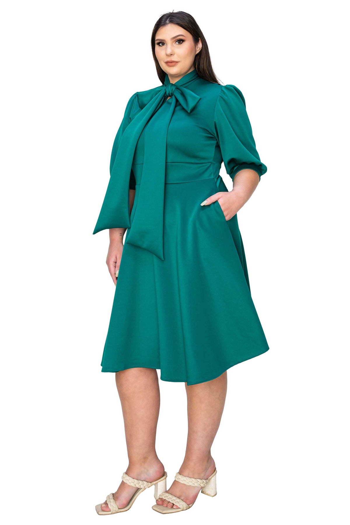A woman modeling a teal green midi dress with a high neck, large bow at the collar, puffed elbow-length sleeves, a fitted waist, and side pockets, paired with beige heeled sandals against a plain white background.
