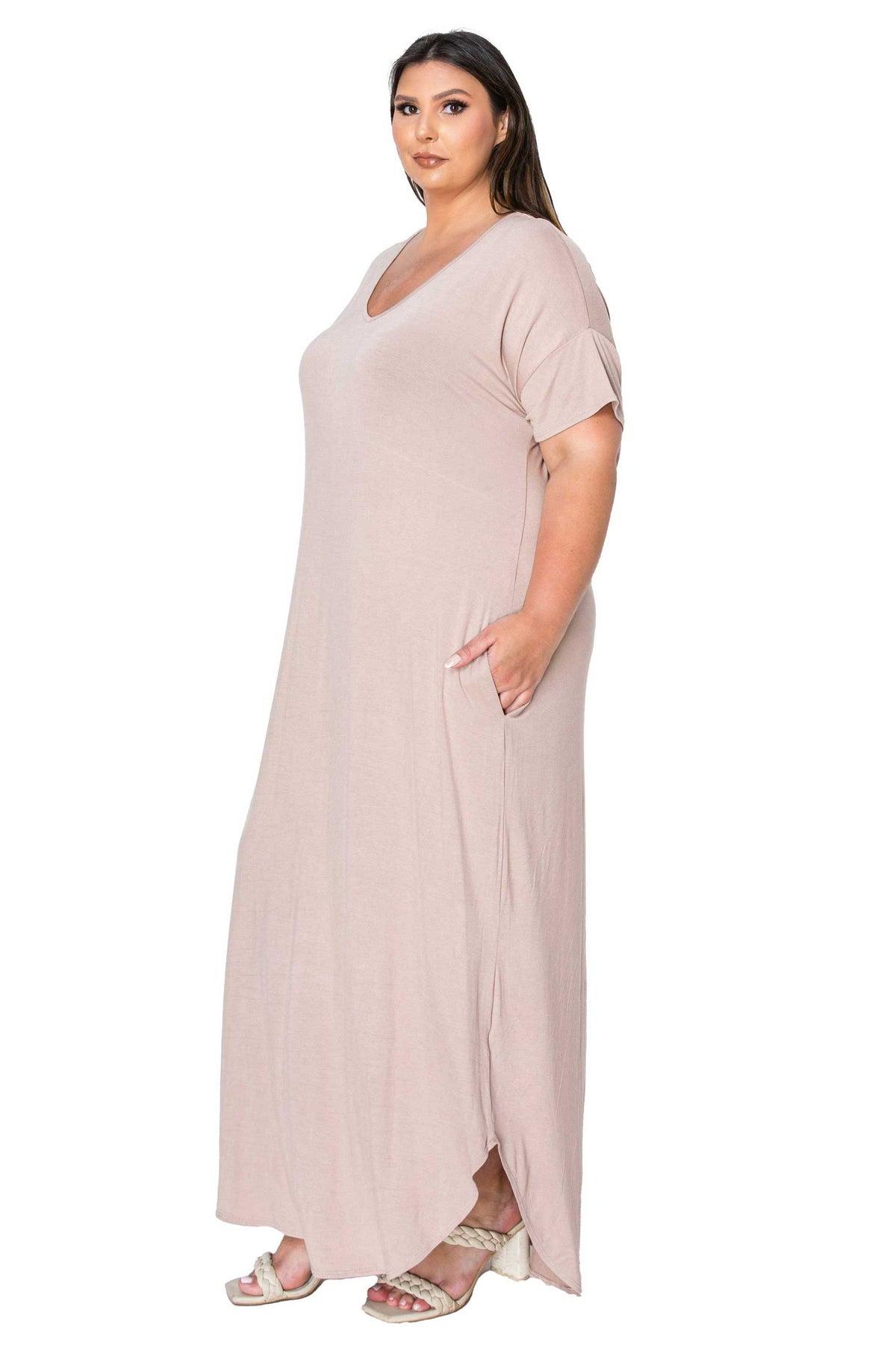 A woman modeling a long, loose-fitting maxi dress in a light blush pink color. The dress features a V-neckline, short sleeves, and side pockets, shown against a plain white background.