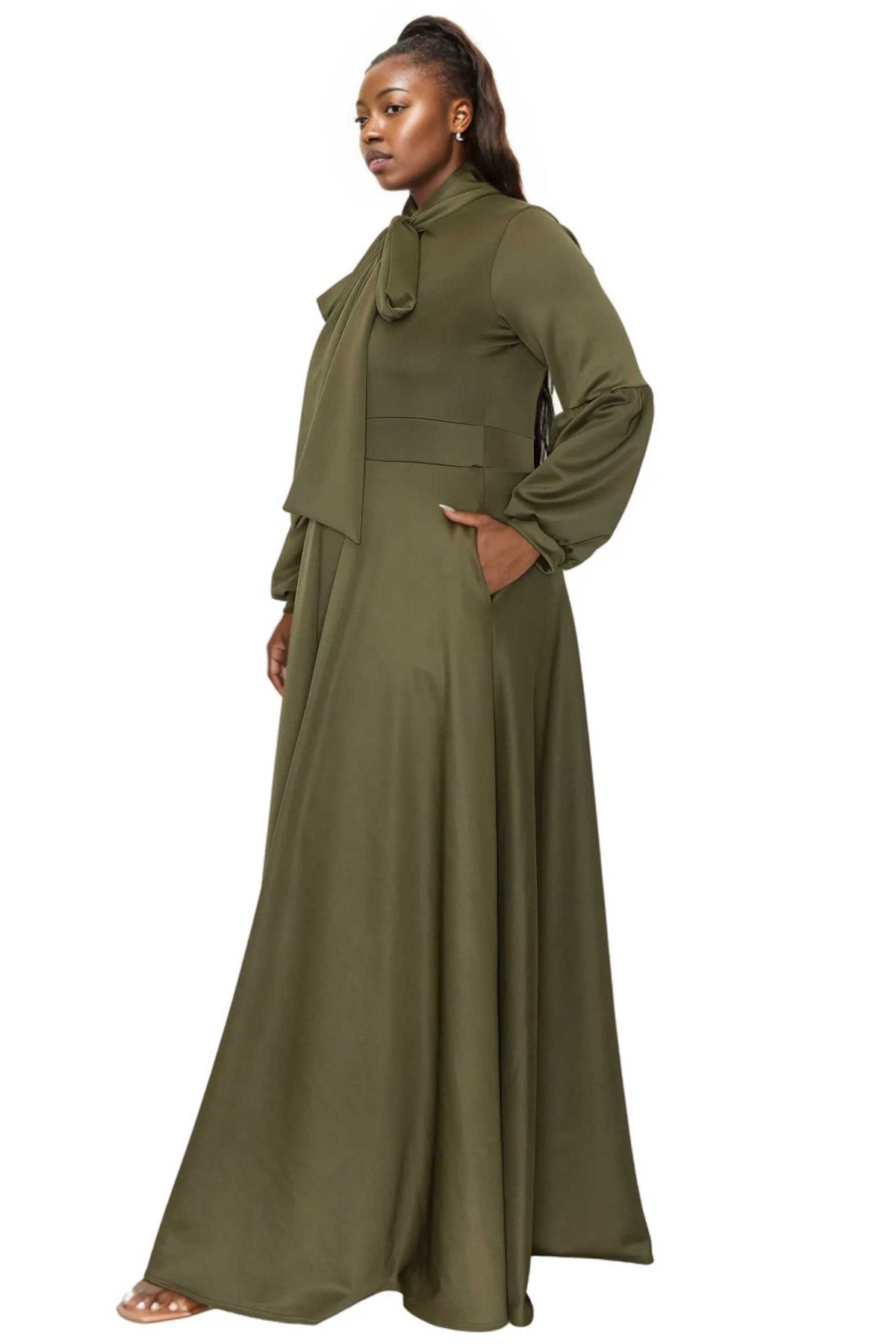 A woman modeling a long-sleeved olive green maxi dress with a high neck, tie detail at the collar, and a matching draped scarf, shown against a plain white background.