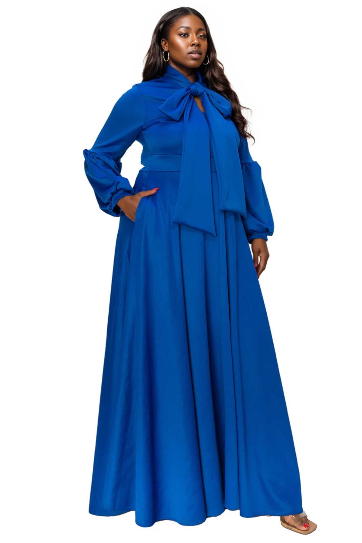 A woman modeling a long-sleeved royal blue maxi dress with a large bow at the neckline, a matching waist tie, and a flowing skirt, set against a plain white background.