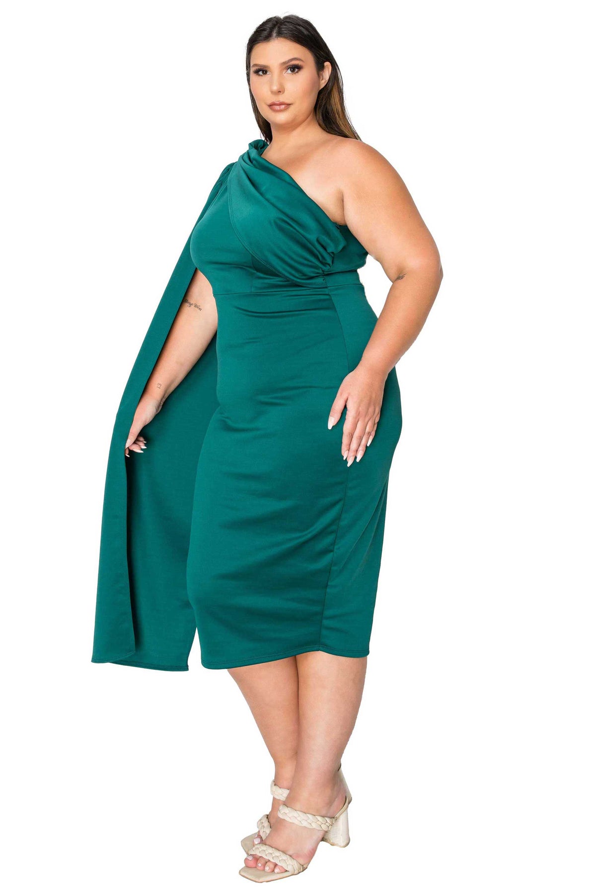 A woman modeling a one-shoulder emerald green midi dress with a draped cape-style sleeve, paired with beige strappy high-heeled sandals, against a plain white background.