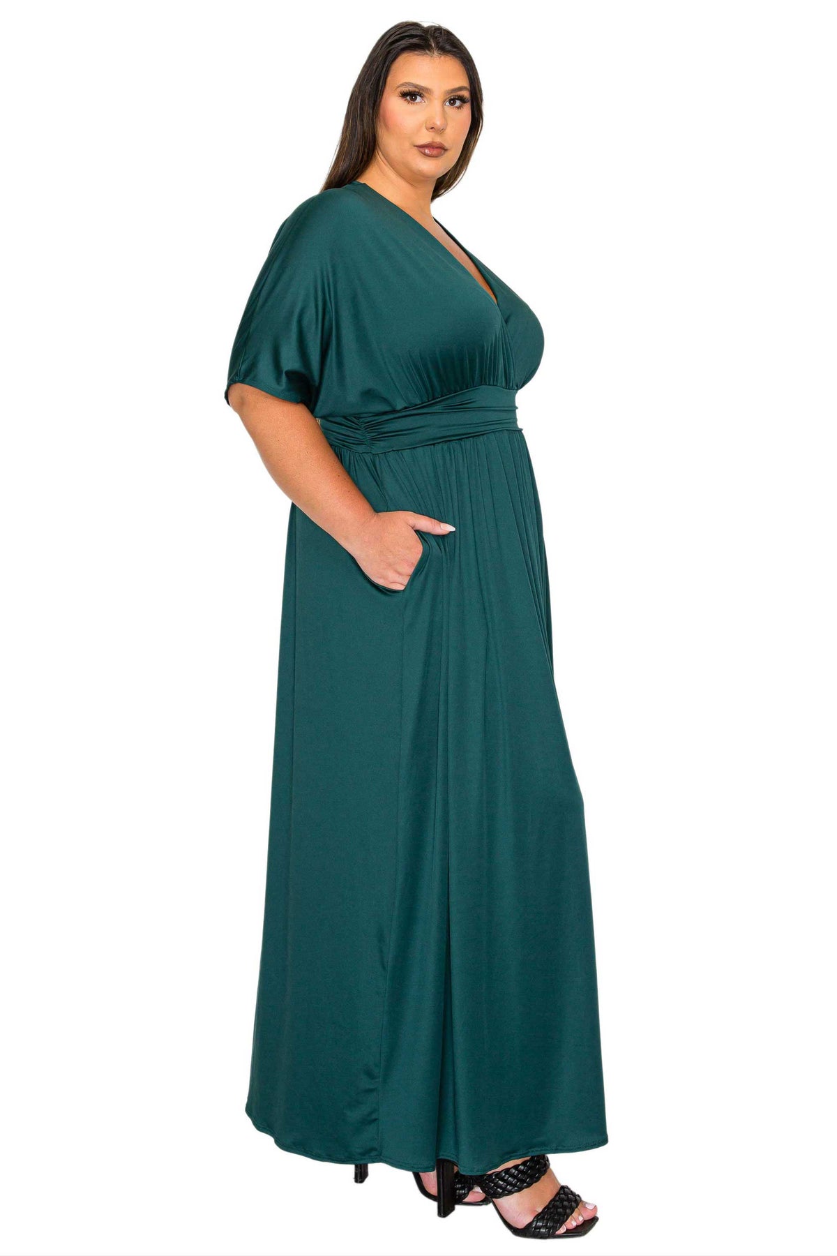 A woman modeling a dark green maxi dress with short flutter sleeves, a V-neckline, and side pockets, paired with black heeled sandals against a plain white background.