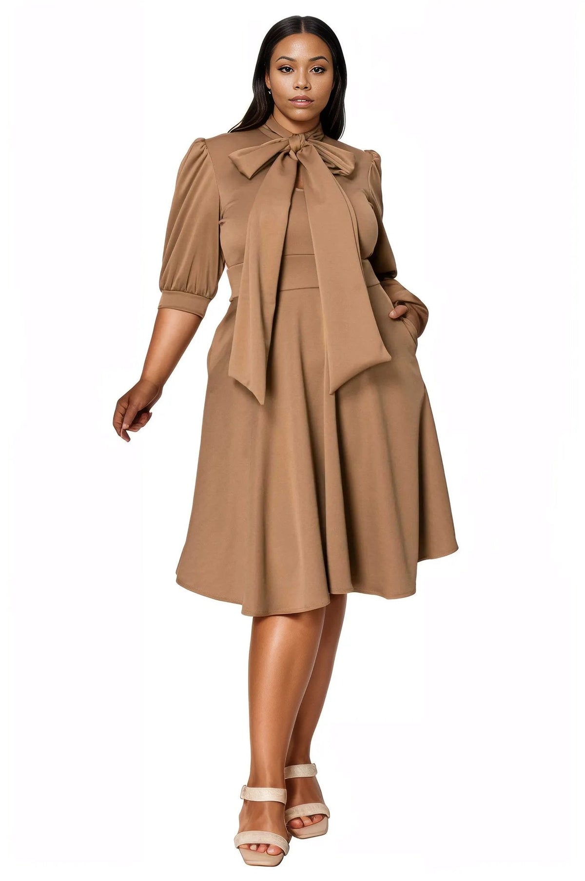 A woman modeling a tan midi dress with puff sleeves, a large bow tie neckline, and a flared skirt, paired with beige heeled sandals against a plain white background.