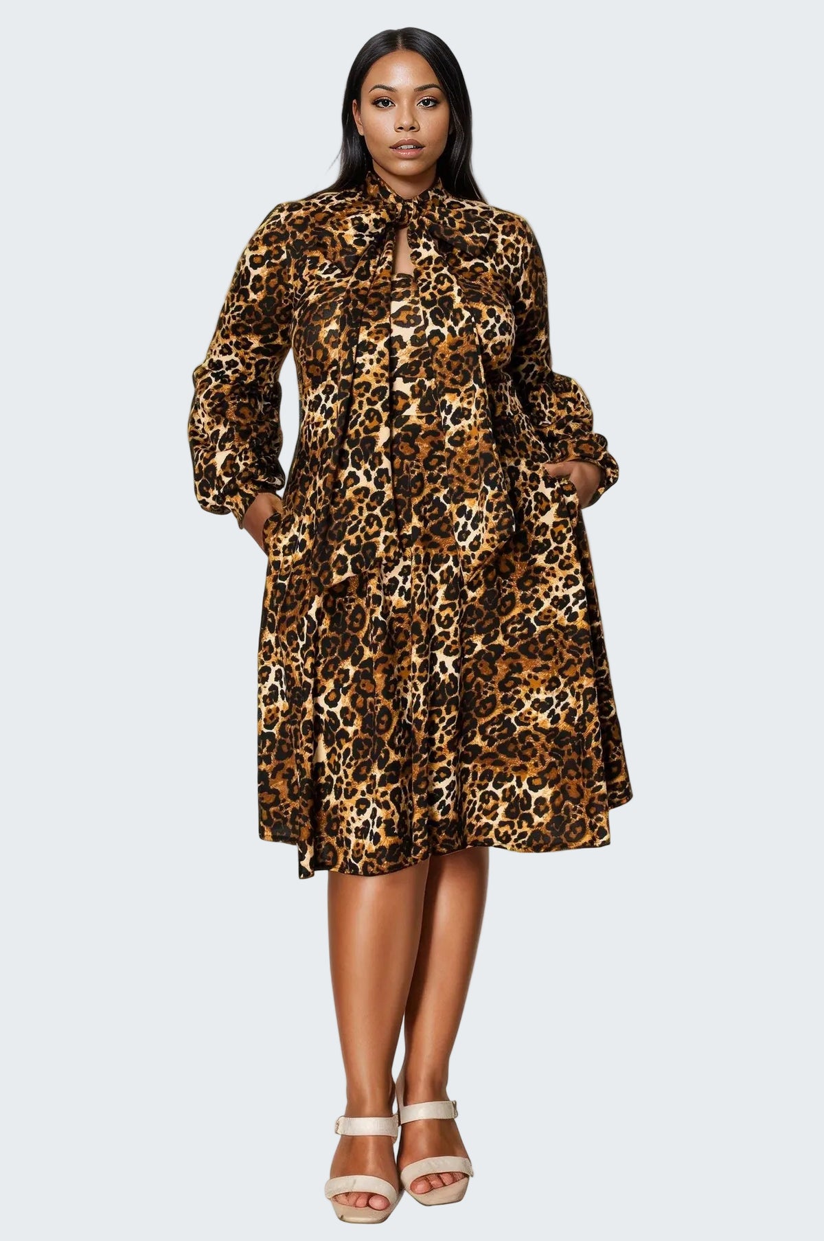 A woman modeling a leopard-print long-sleeve midi dress with a tie-neck and front pockets, paired with beige platform sandals, against a plain white background.
