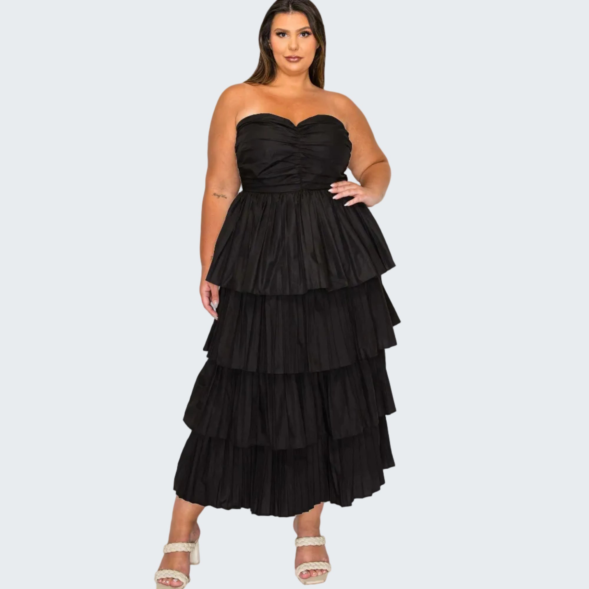A woman modeling a strapless black maxi dress with a ruched sweetheart bodice and a tiered, ruffled skirt, paired with white heeled sandals against a plain white background.