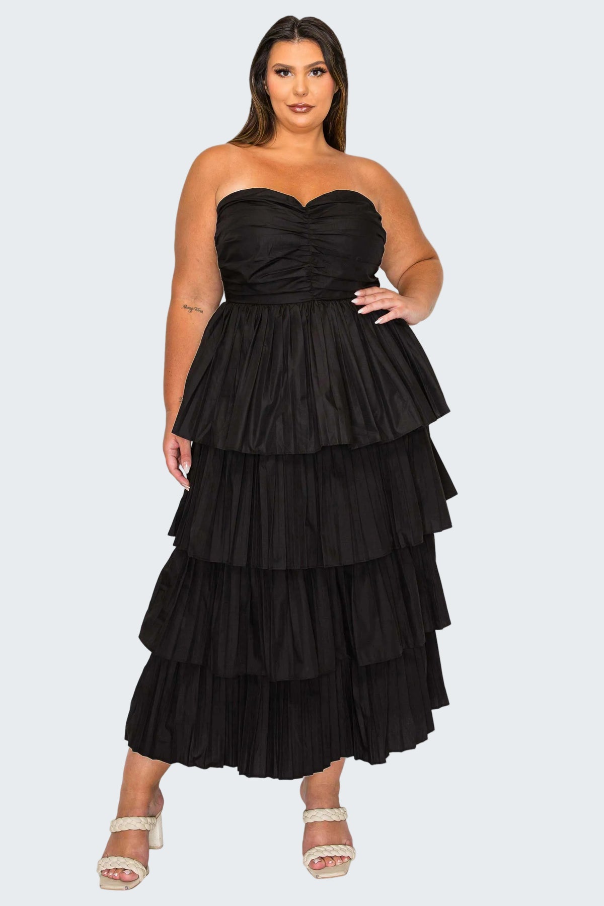 A woman modeling a strapless black maxi dress with a ruched sweetheart bodice and a tiered, ruffled skirt, paired with white heeled sandals against a plain white background.