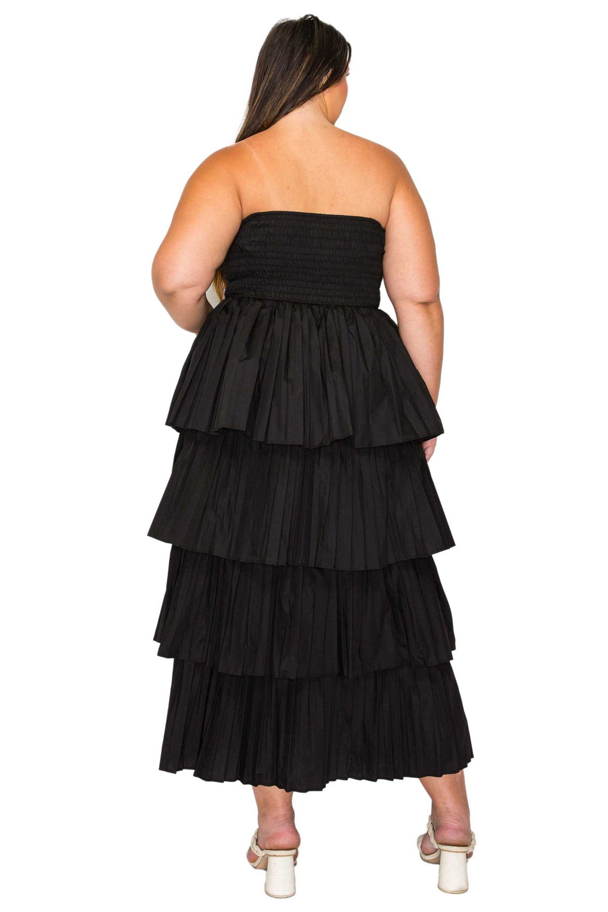 A woman shown from the back wearing a strapless black tiered maxi dress with white block-heel sandals, set against a plain white background.