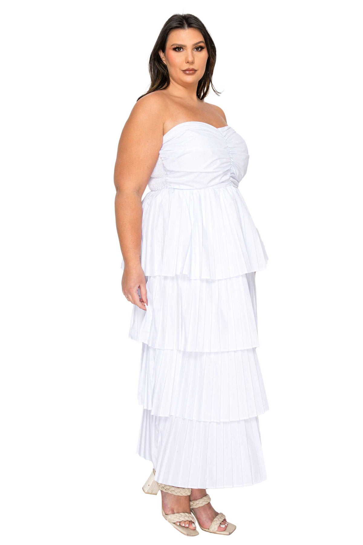 A woman modeling a white strapless tiered maxi dress with a ruched bodice and pleated skirt, paired with beige strappy heels, against a plain white background.