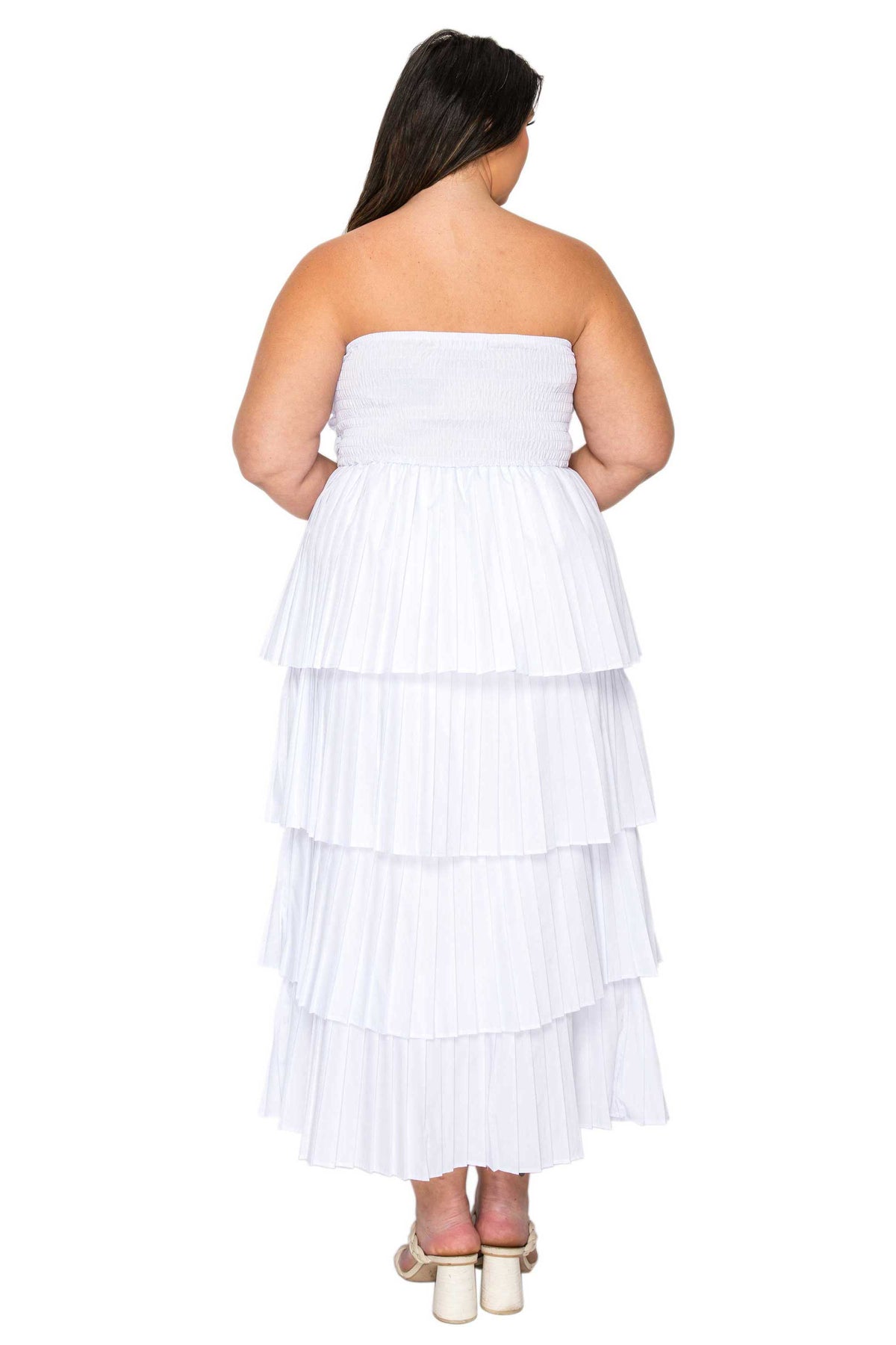 A woman viewed from the back wearing a strapless white tiered maxi dress with a pleated ruffled skirt and white block-heel sandals, set against a plain white background.