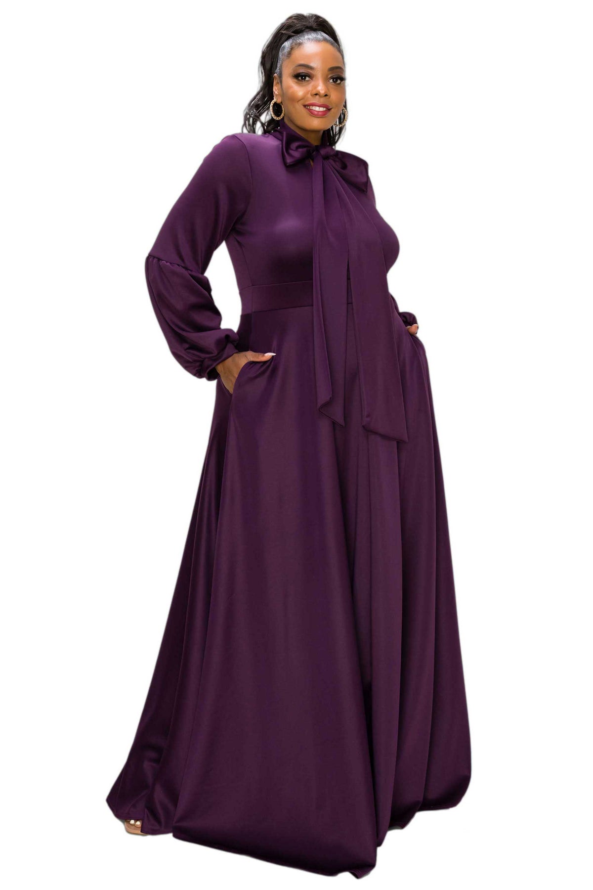 A woman modeling a long-sleeved, floor-length purple maxi dress with a high neck, bow detail at the collar, and side pockets, set against a plain white background.