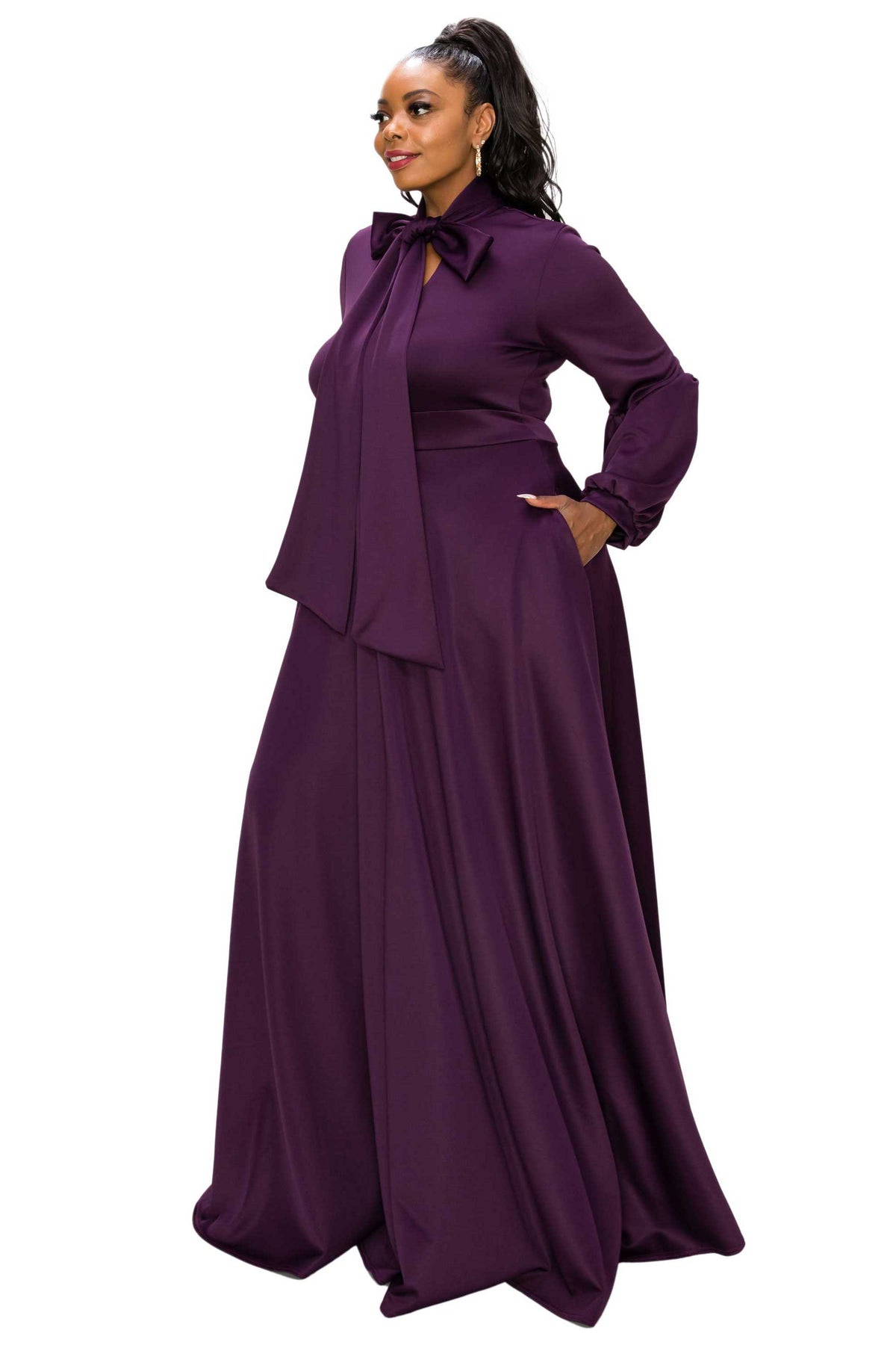 A woman modeling a long-sleeved, floor-length purple gown with a high neck, bow detail at the collar, and a draped overlay, set against a plain white background.