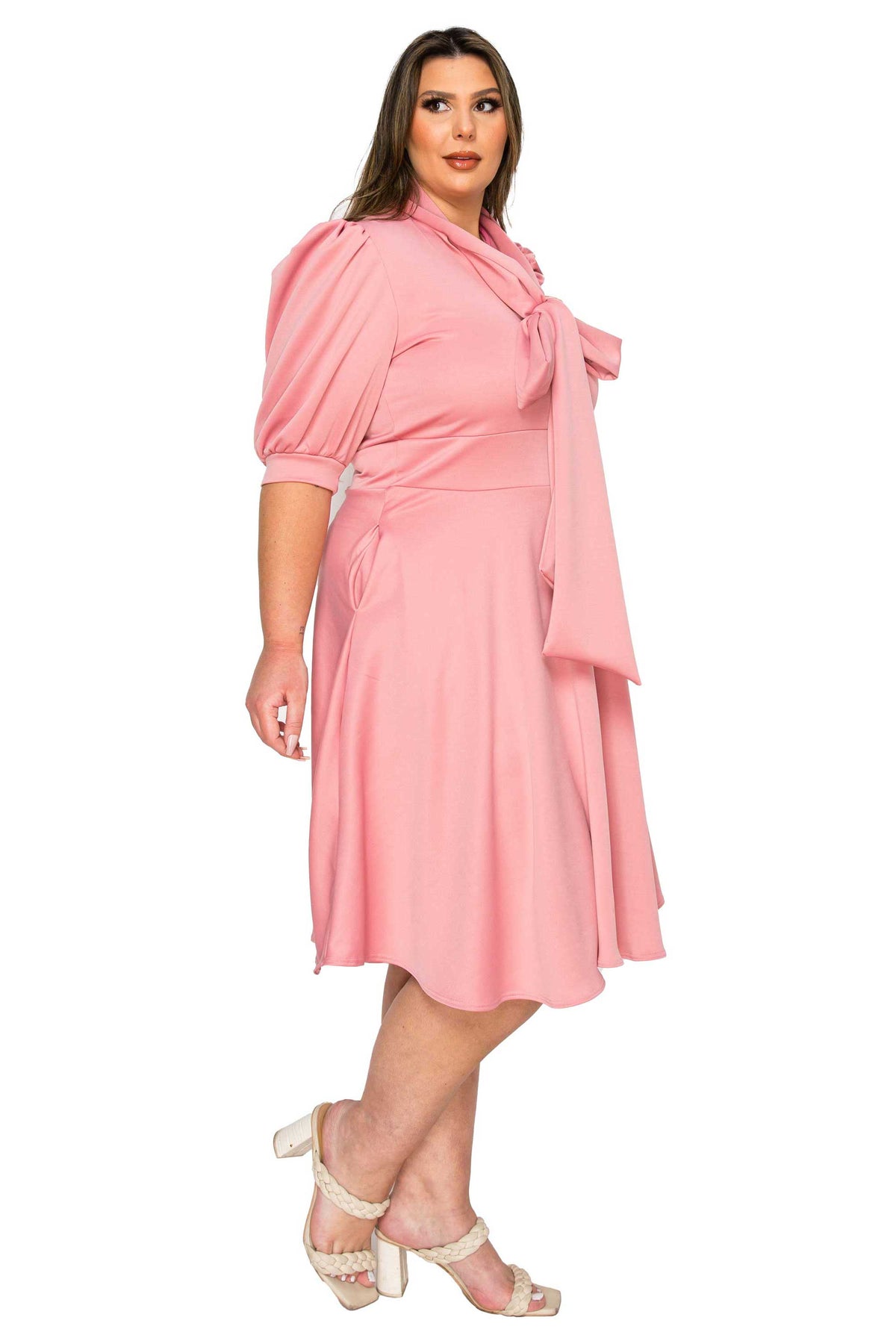 A woman modeling a light pink midi dress with puffed elbow-length sleeves, a wrap-style tie neckline, and a flared skirt, paired with cream high-heeled sandals against a plain white background.