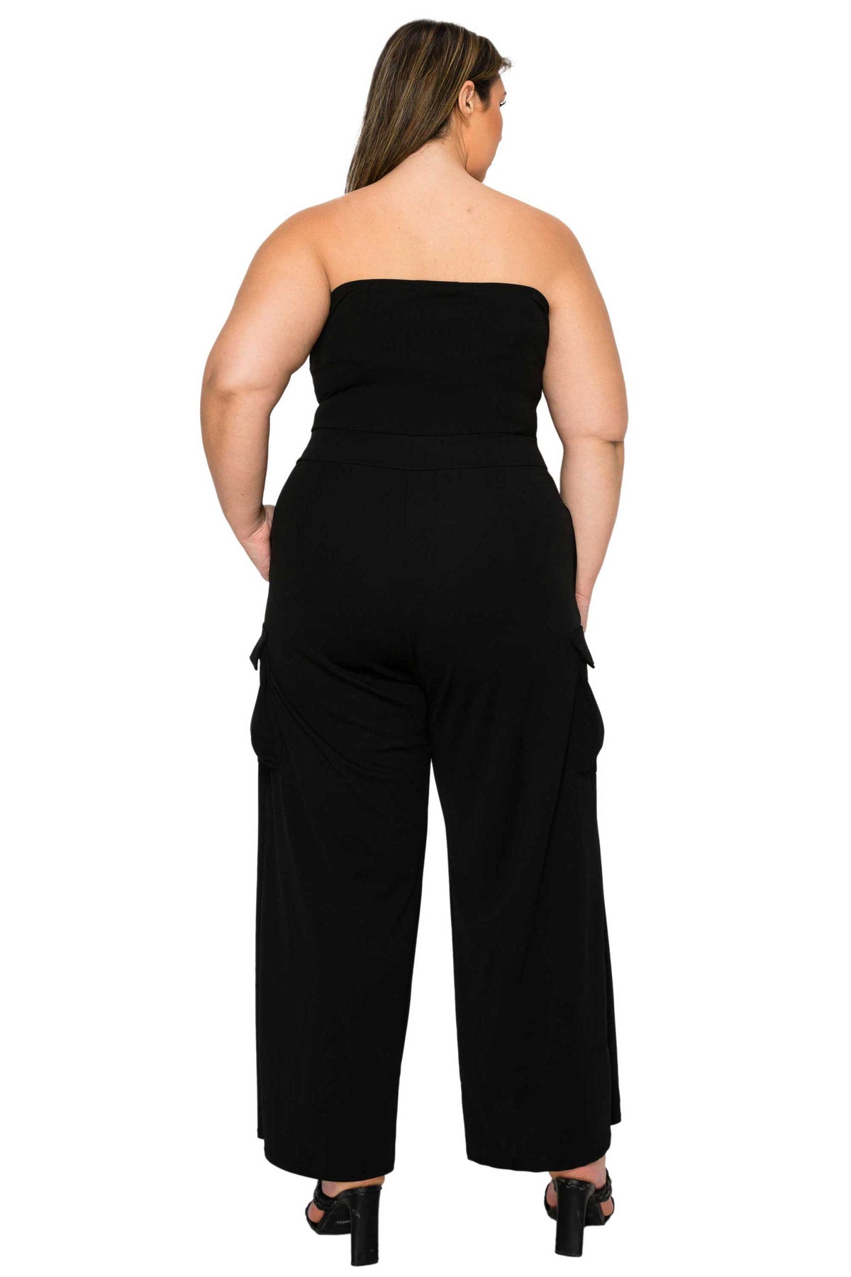 A plus-size model viewed from the back wearing a black strapless wide-leg jumpsuit with cargo pockets, paired with black high-heeled sandals, against a plain white background.