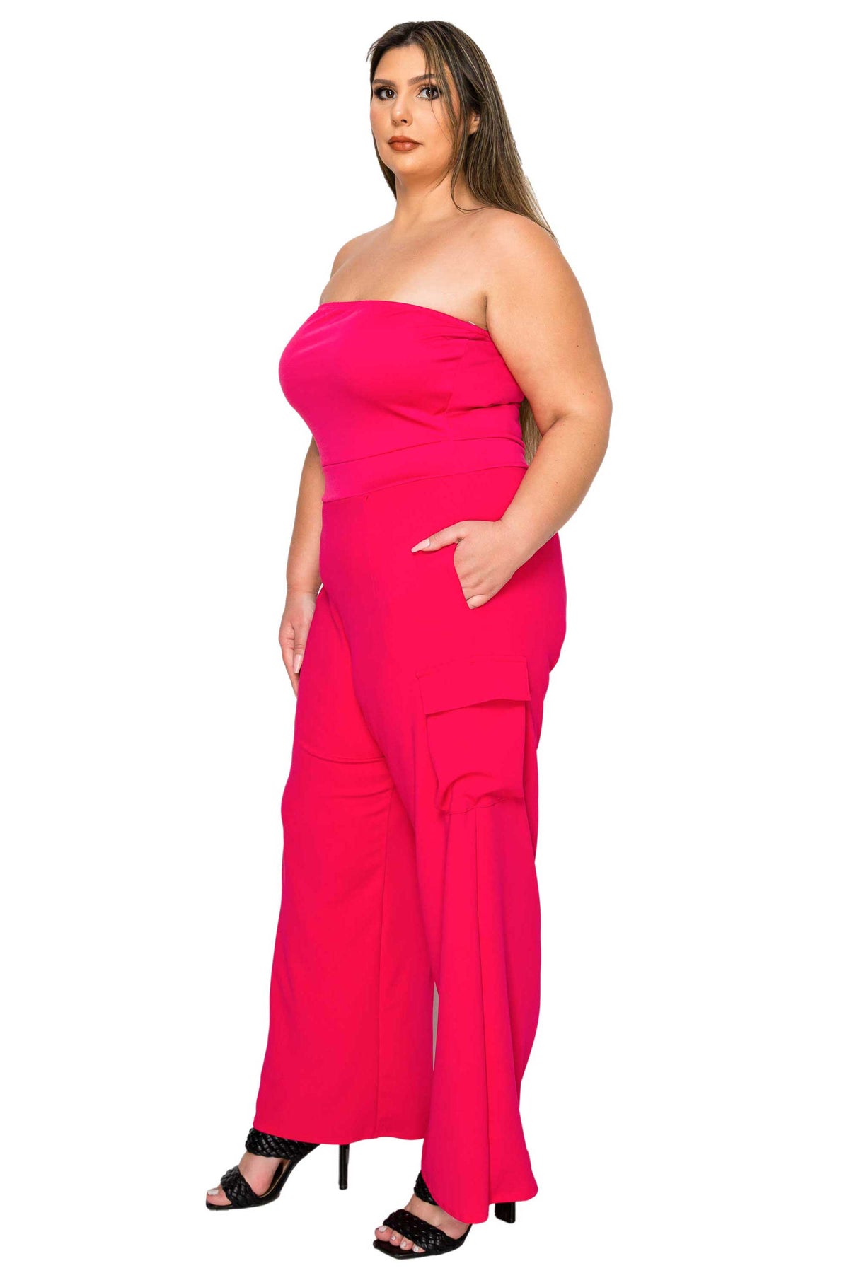A woman modeling a bright hot pink strapless jumpsuit with wide legs and side pockets, paired with black heeled sandals, against a plain white background.