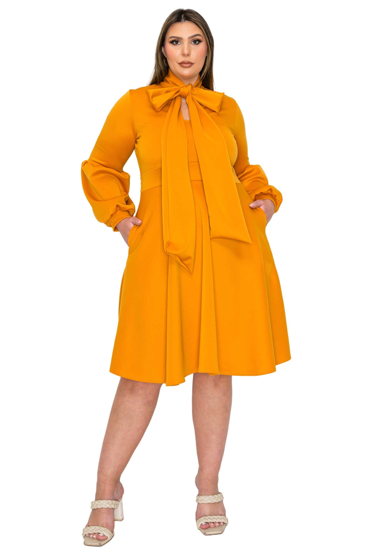 A woman modeling a bright yellow-orange long-sleeve midi dress with a large bow tie-neck detail, side pockets, and a flared skirt, paired with white heeled sandals against a plain white background.