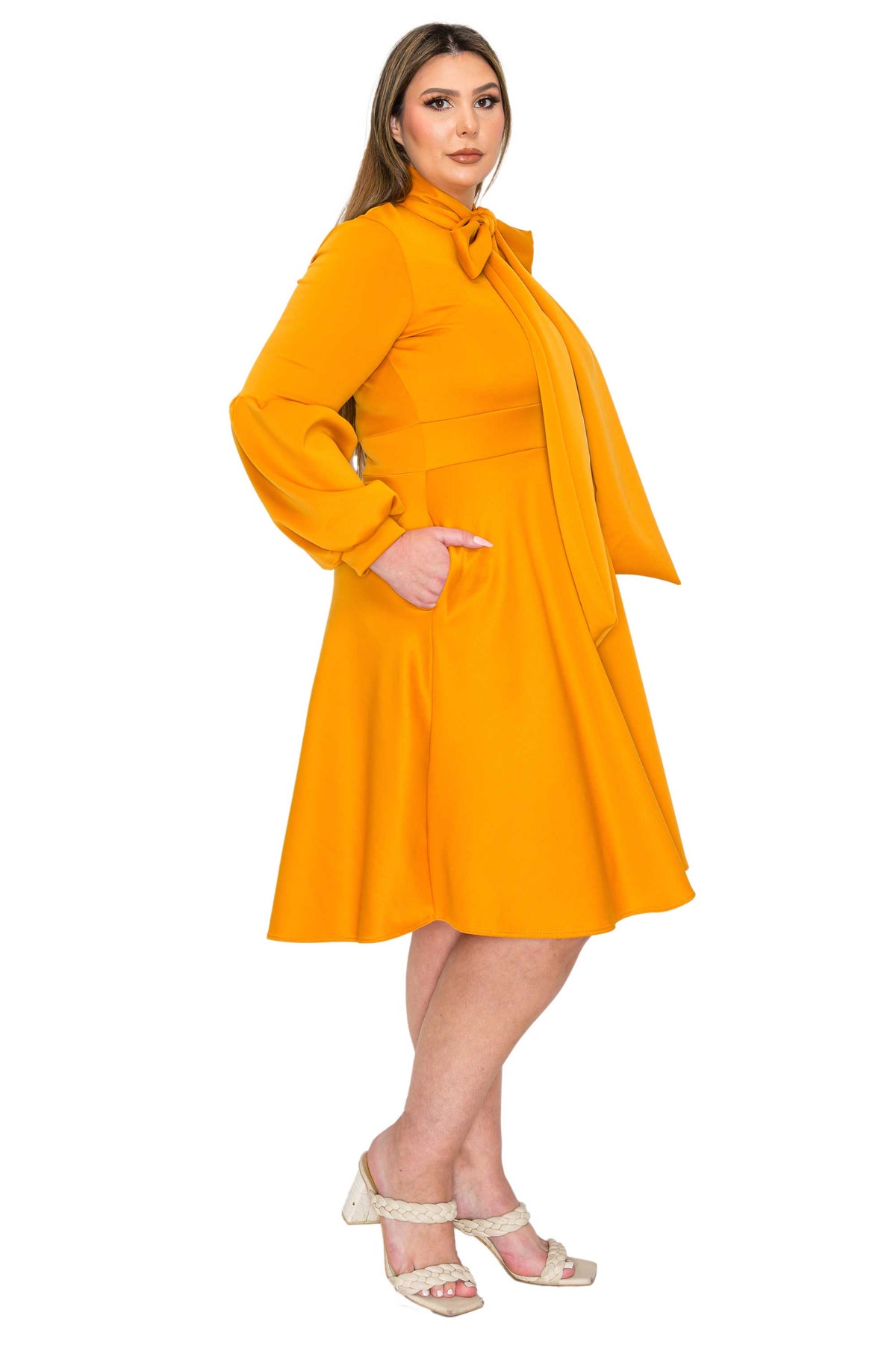A woman modeling a bright yellow-orange long-sleeve dress with a high collar, wide lapels, and side pockets, paired with beige strappy heels, against a plain white background.