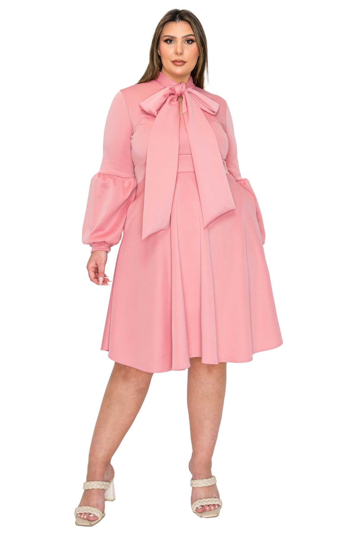 A woman modeling a pink long-sleeve midi dress with a large bow at the neckline, puff sleeves, and a flared skirt, paired with white heeled sandals against a plain white background.