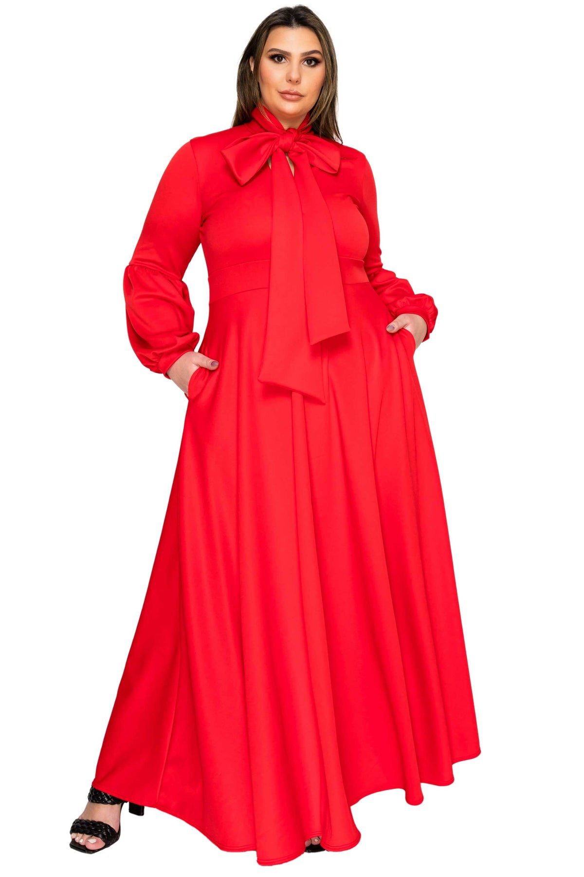 A woman modeling a bright red long-sleeve maxi dress with a large bow at the collar, side pockets, and a flowing skirt, photographed against a plain white background.